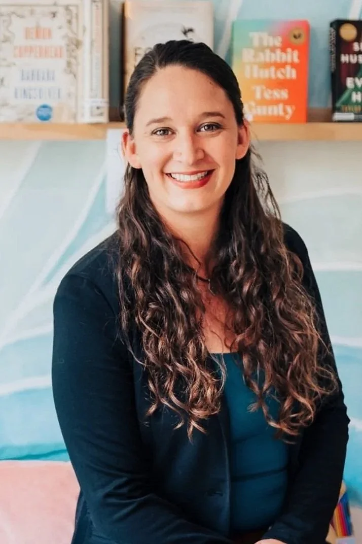 Female therapist with long wavy brown hair wearing a dark jacket and a colorful blue background