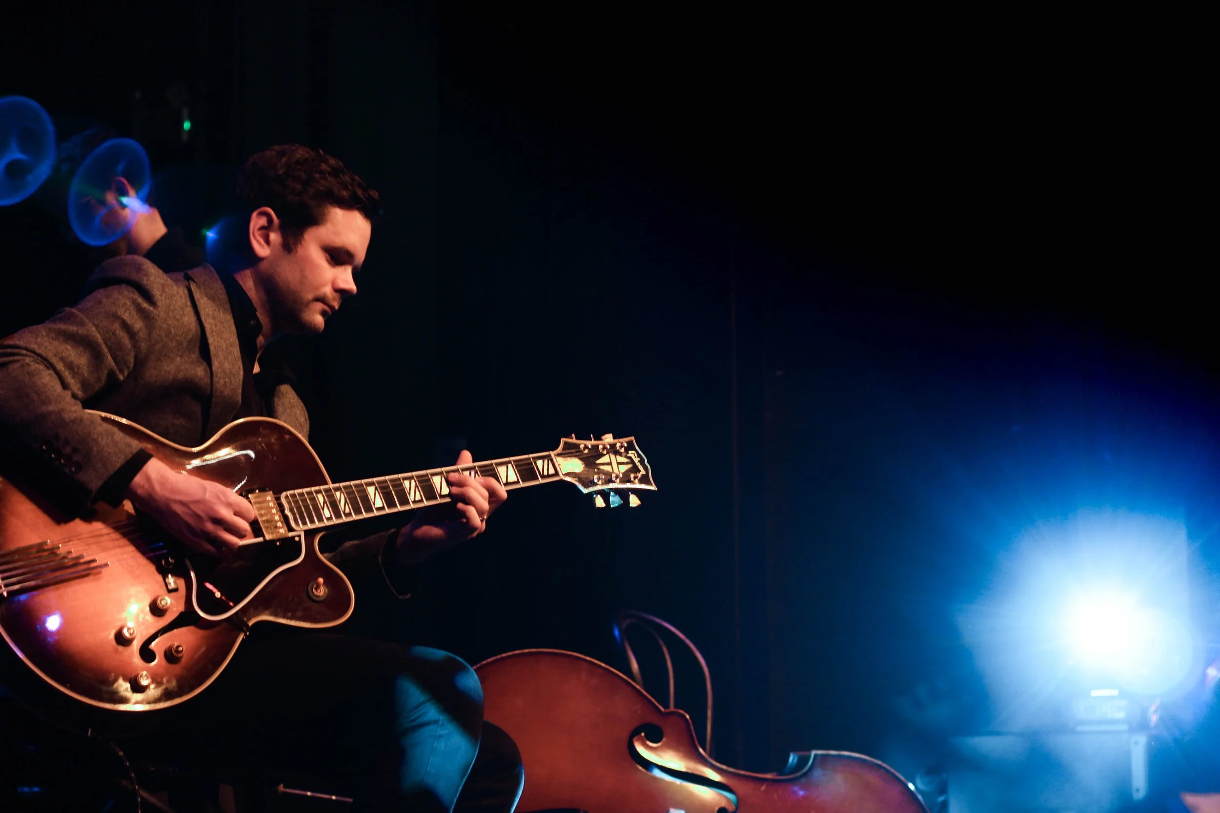 Man playing a guitar on stage with blue lighting and a cello in the background.