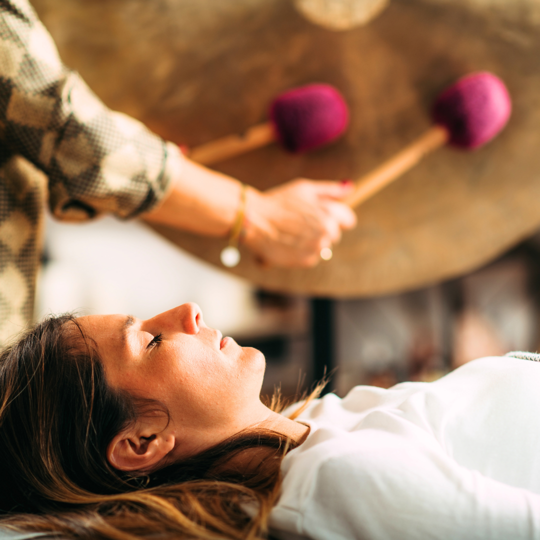 En kvinne ligger med lukket øyne mens en annen person spiller på et slagverkinstrument med to kuler av fiolett ull.