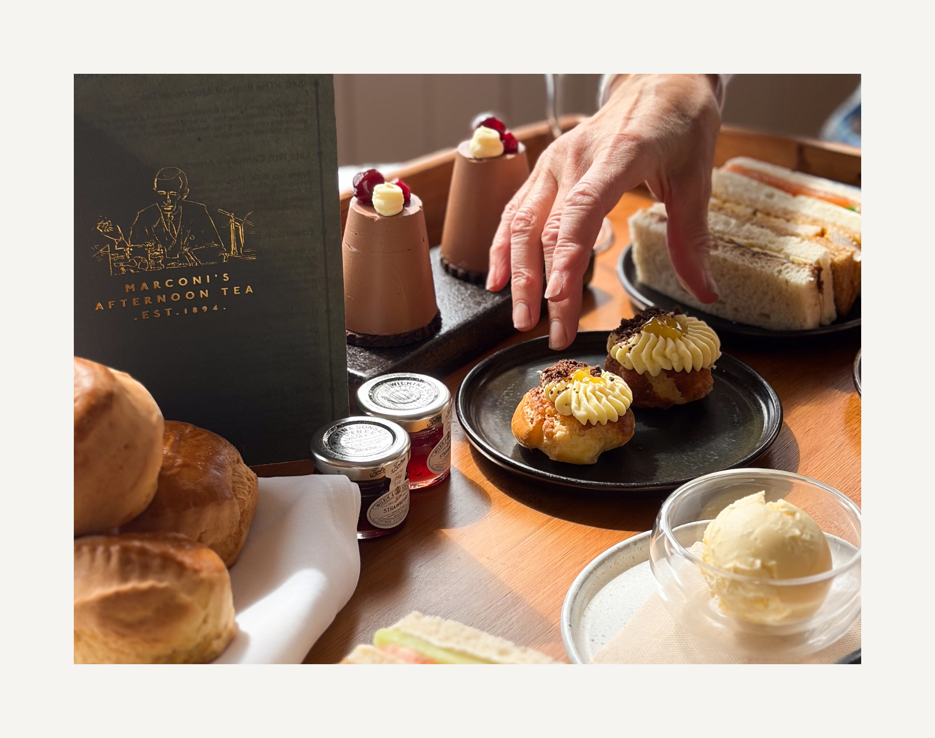 A variety of desserts including cupcakes with cream and cherries, a bowl of vanilla ice cream, and small jars of jam or preserves on a table, with a person's hand reaching for a cupcake.