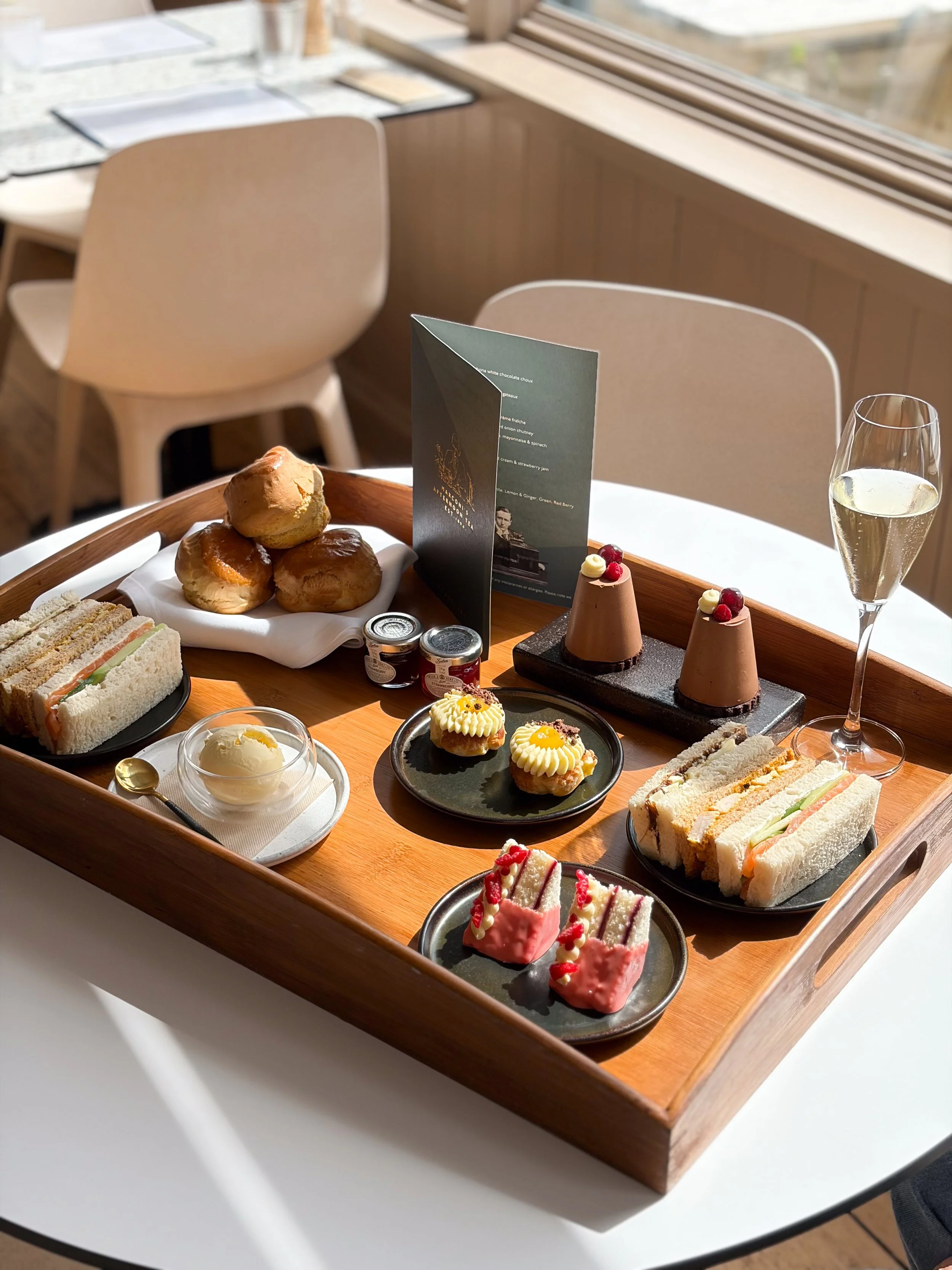 A wooden serving tray with assorted pastries and desserts on a white table near a window, including sandwiches, scones, cupcakes, macarons, and a glass of white wine.