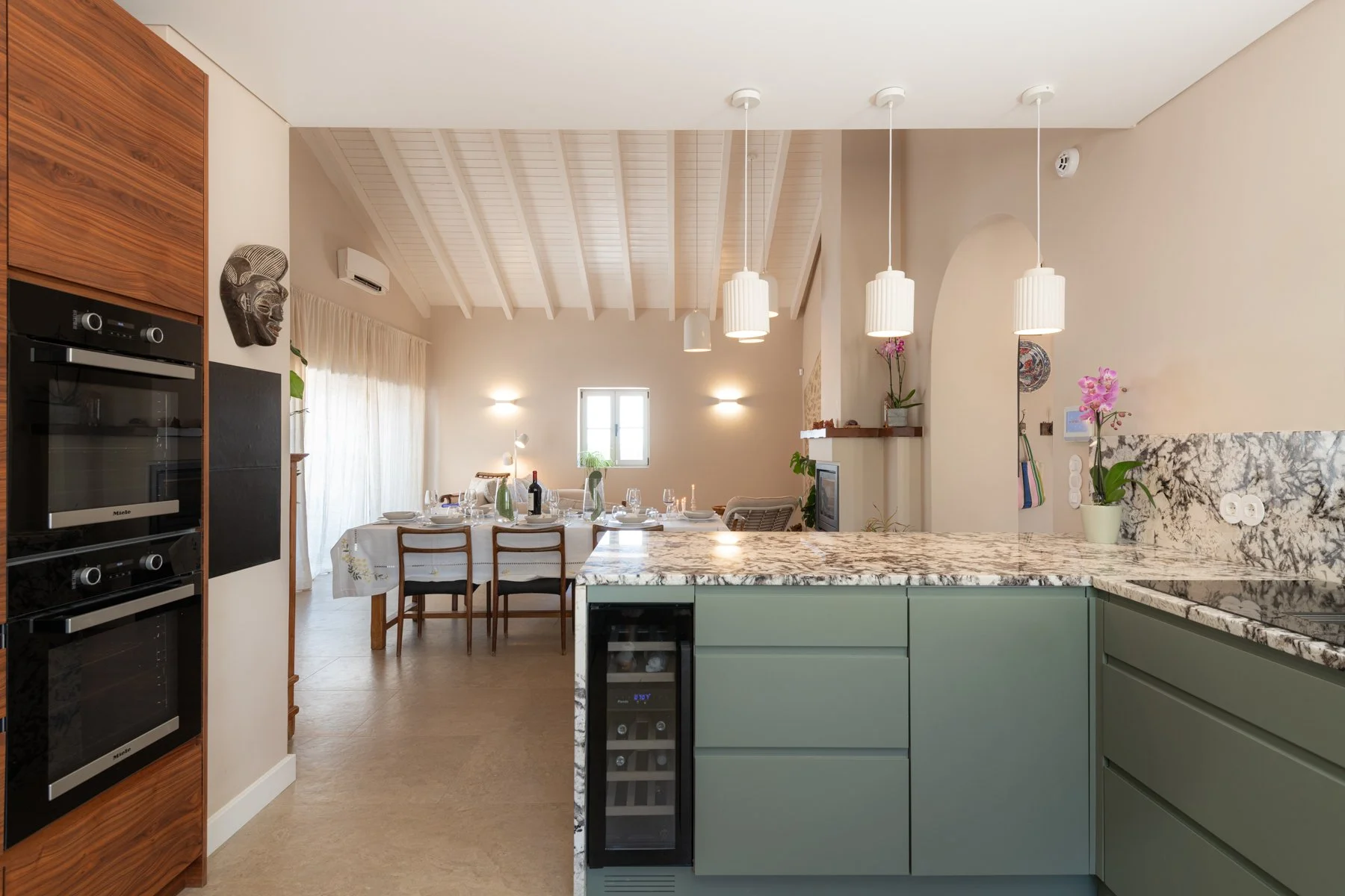 Open-concept kitchen and dining area with vaulted ceiling, hanging pendant lights, green cabinets, marble countertops, potted orchids, and a dining table set for a meal.