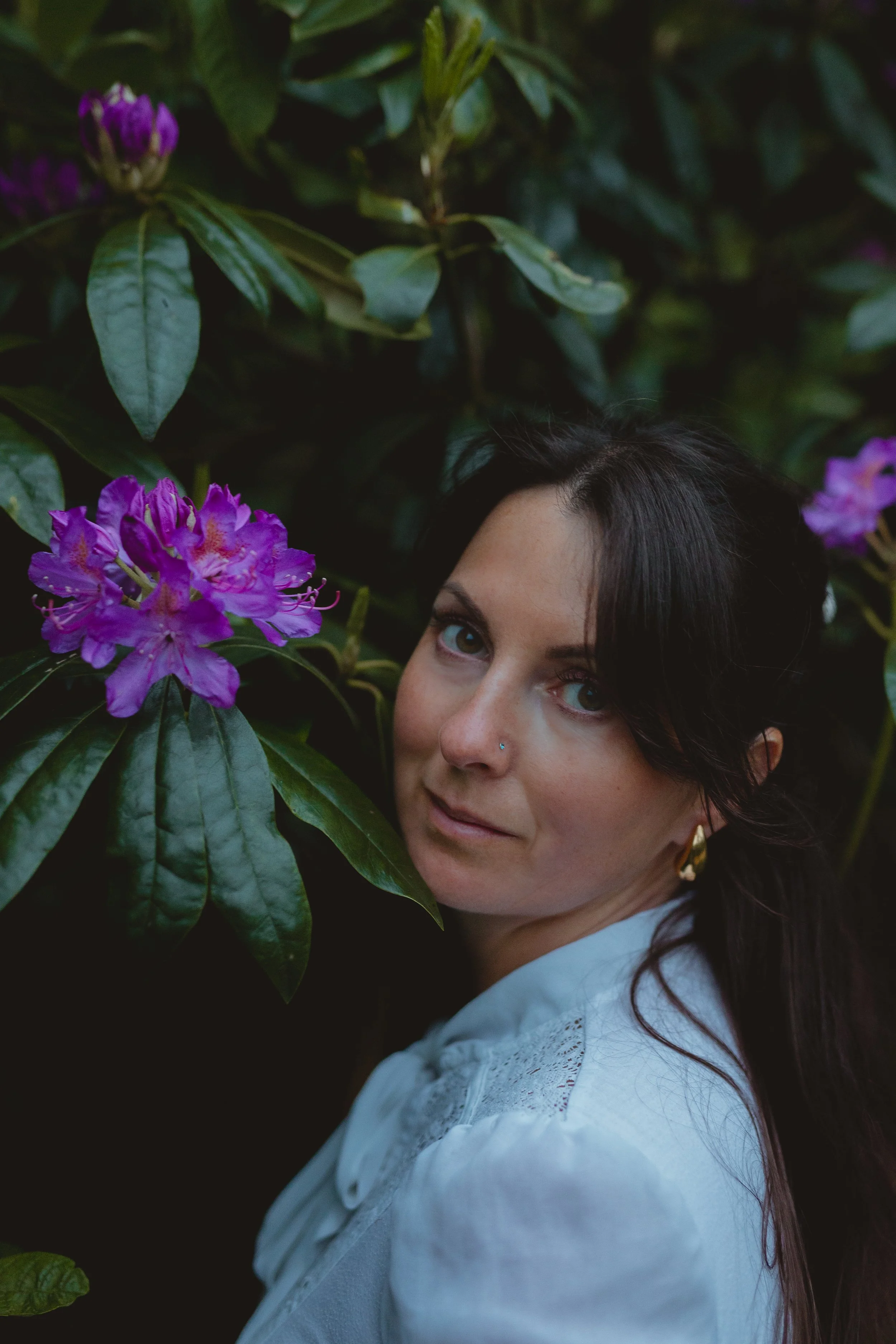 Portrait of a holistic woman with dark hair and in a white blouse with gold earrings looking at the camera with a green bush with bright purple flowers behind her in Somerleyton Suffolk 