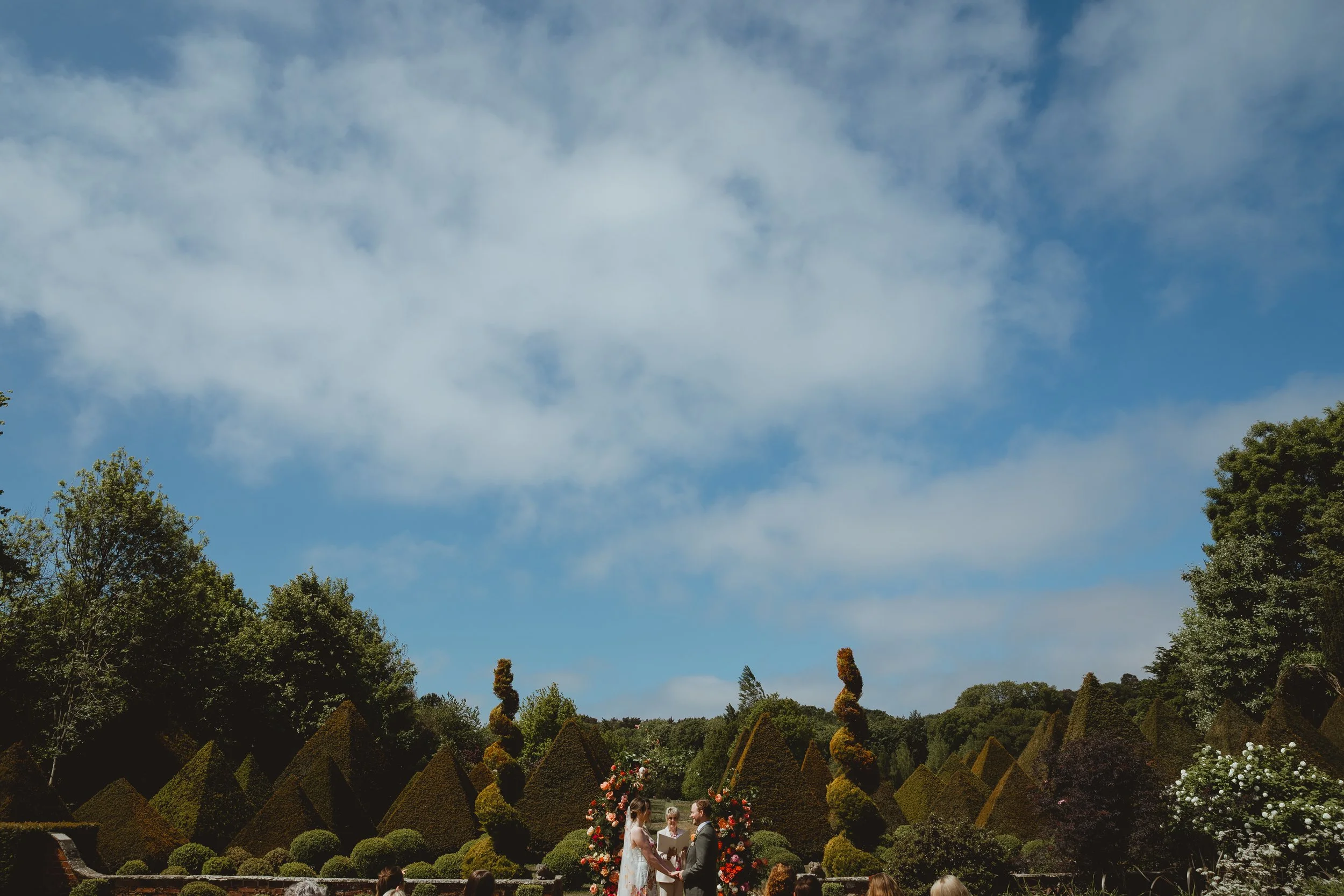 Wide shot of an outdoor wedding ceremony set in a formal garden with sculpted hedges under a blue sky at Chaucer Barn Gresham Holt Norfolk 