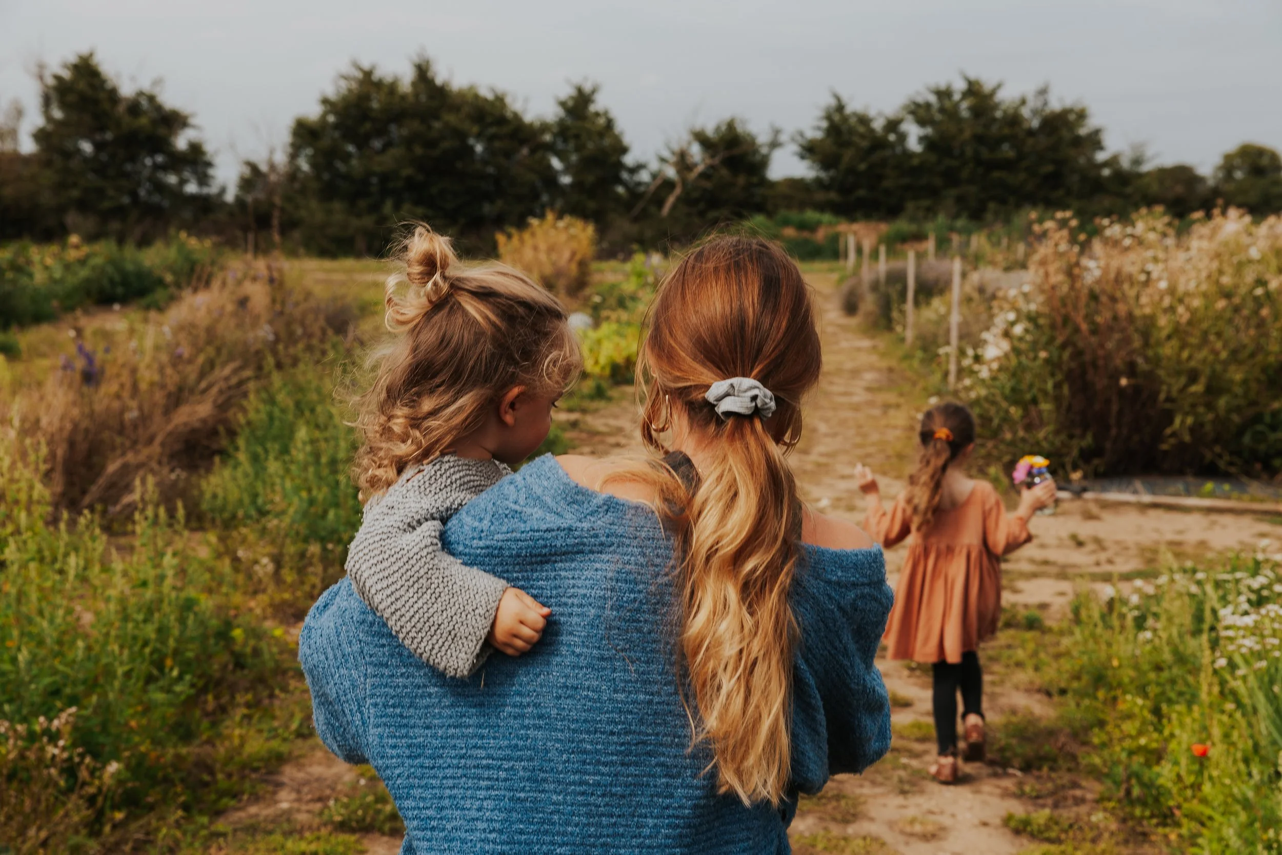 Wide shot of a blonde mother from the back in a blue outfit whilst her son and daughter run ahead of her in a green flower field in Suffolk Southwold 
