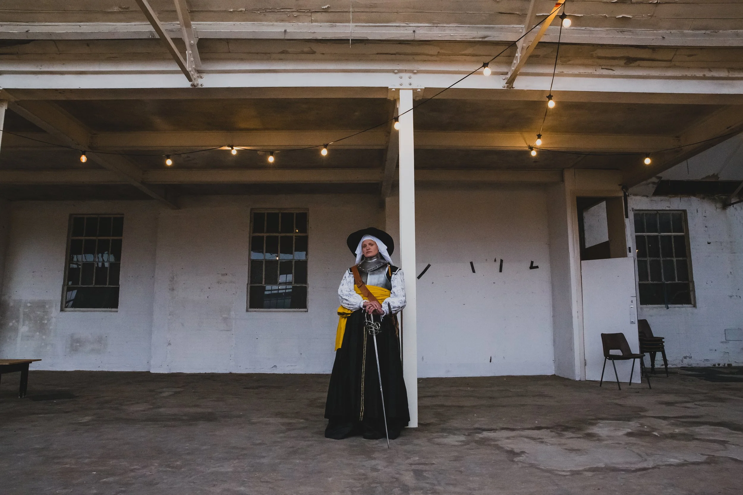 Editorial wide shot taken in a factory in Norwich Norfolk of a strong woman dressed in medieval fighting attire with a sword and festoon lights in the foreground 