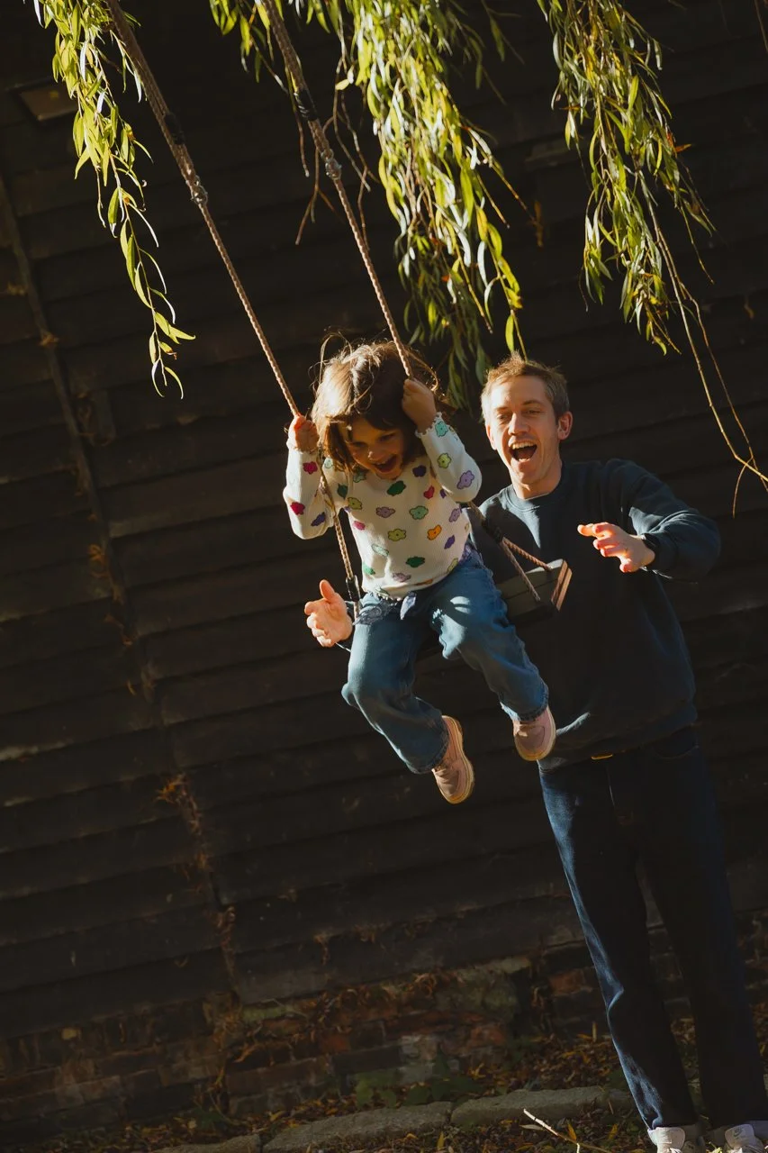 Dark cinematic shot of a father pushing his daughter on a swing lit up by the sun with green leaves hanging in the foreground at Restaries Halesworth Suffolk 