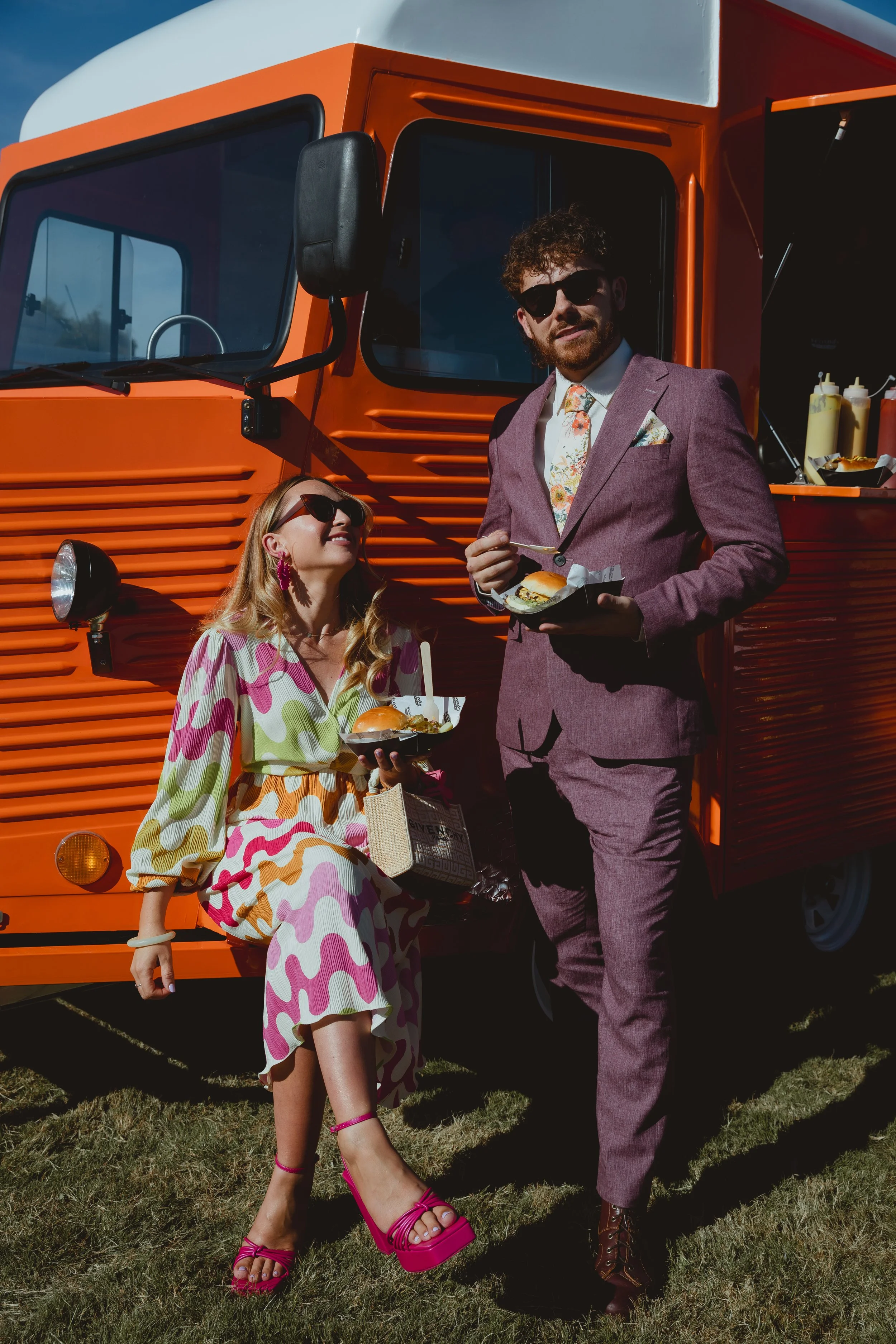 Bright vibrant documentary image of two wedding guests vibrantly dressed in patterned pink orange and purple outfits looking retro sat against a retro food van at an outdoor festival wedding in Blundeston Lowestoft Suffolk 