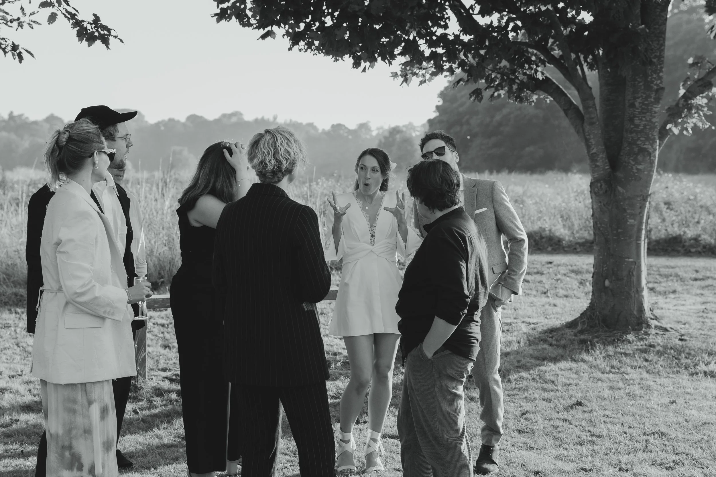 Black and white documentary wedding guest photograph with bride in minidress and bandage heels making a funny face as she talks to friends outside at Sudbourne Hall Orford Suffolk Coast 
