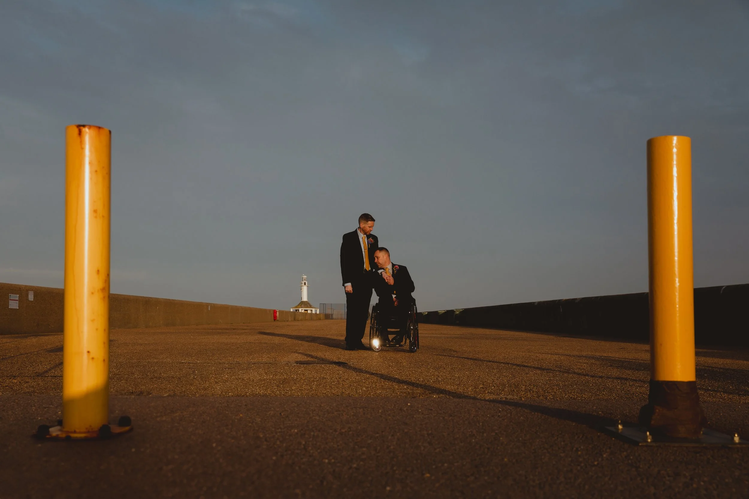 Same sex male groom couple one in a wheelchair hold and caress each other in an editorial wideshot after their wedding at East Point Pavilion on Lowestoft Pier Beach Suffolk Coast