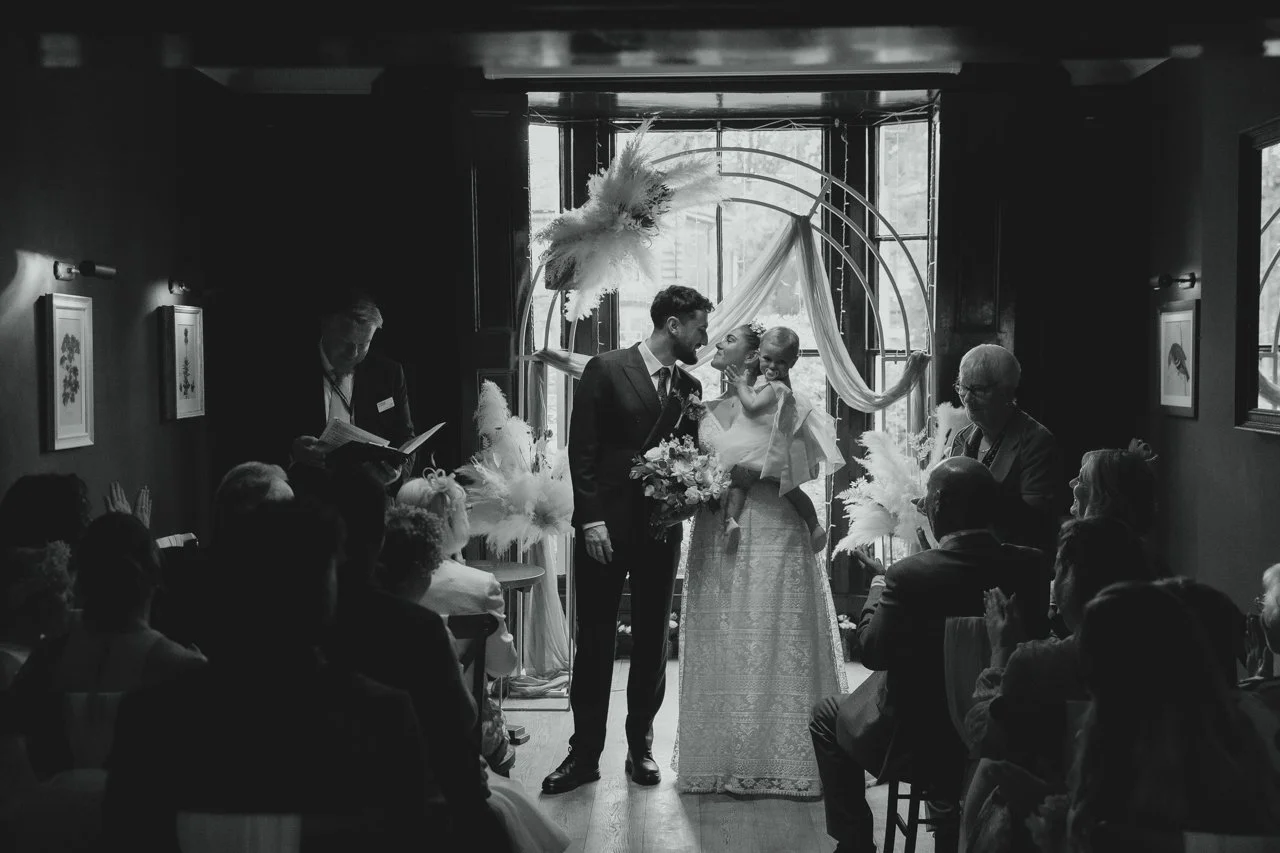 Dark highly contrasted cinematic black and white backlit photograph of bride in a Lavender and Jude dress and groom just married in sophisticated outfits with flower girl at Georgian Townhouse Norwich 