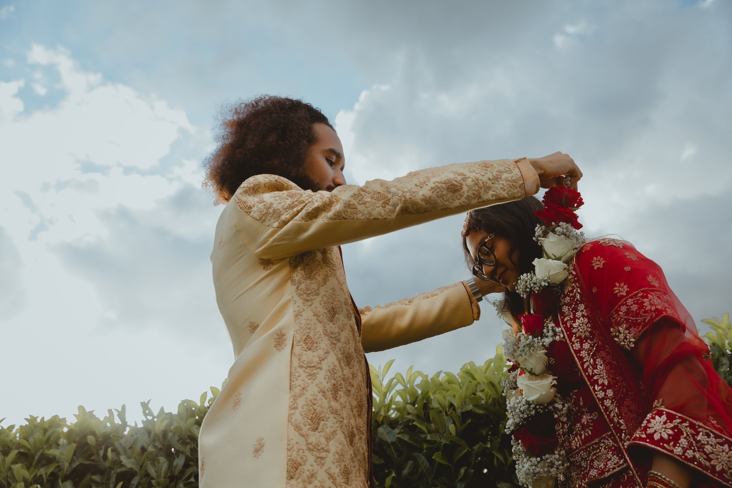 South Asian wedding with mixed race couple putting a flower garland on the bride against a blue sky at Wensum Valley Hotel and Golf Norwich Norfolk 