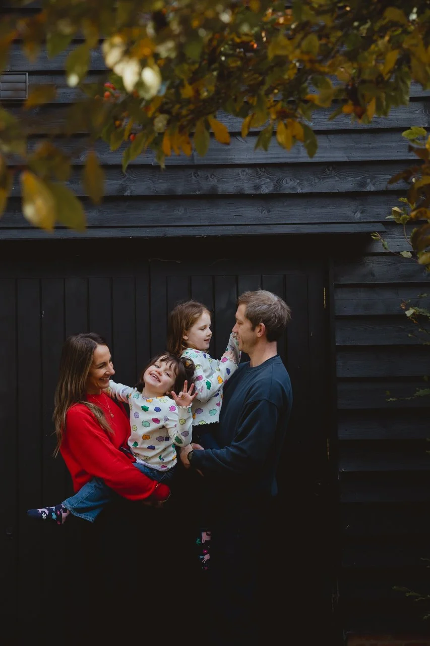 Mum and dad with two daughters against a black barn in bright outfits with golden foliage in the foreground at Restaries Halesworth Suffolk 