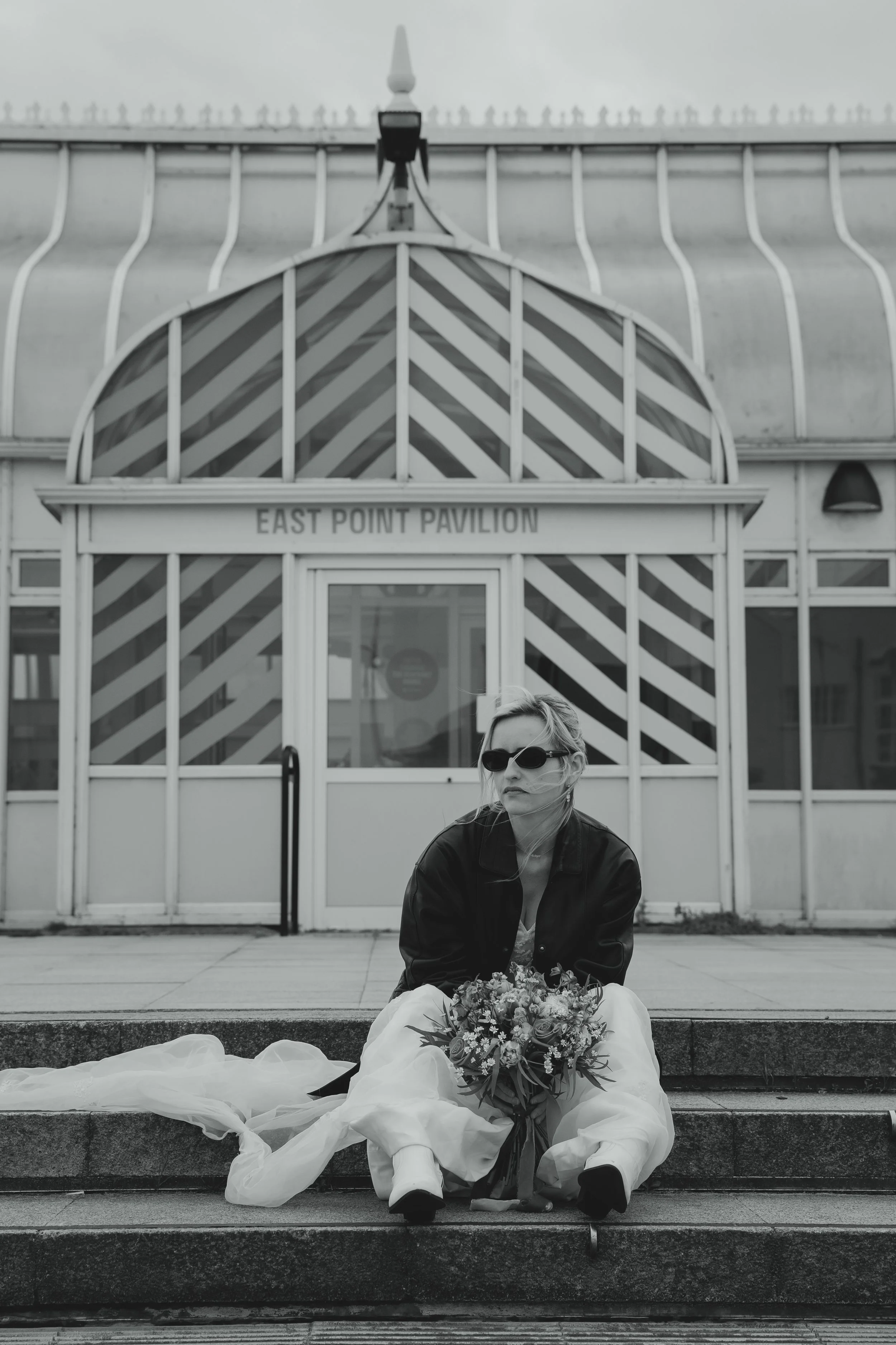 Editorial black and white high contrast image of alternative bride sat with boots leather jacket and sunglasses outside of the East Point Pavilion by the seaside coast in Lowestoft Suffolk 