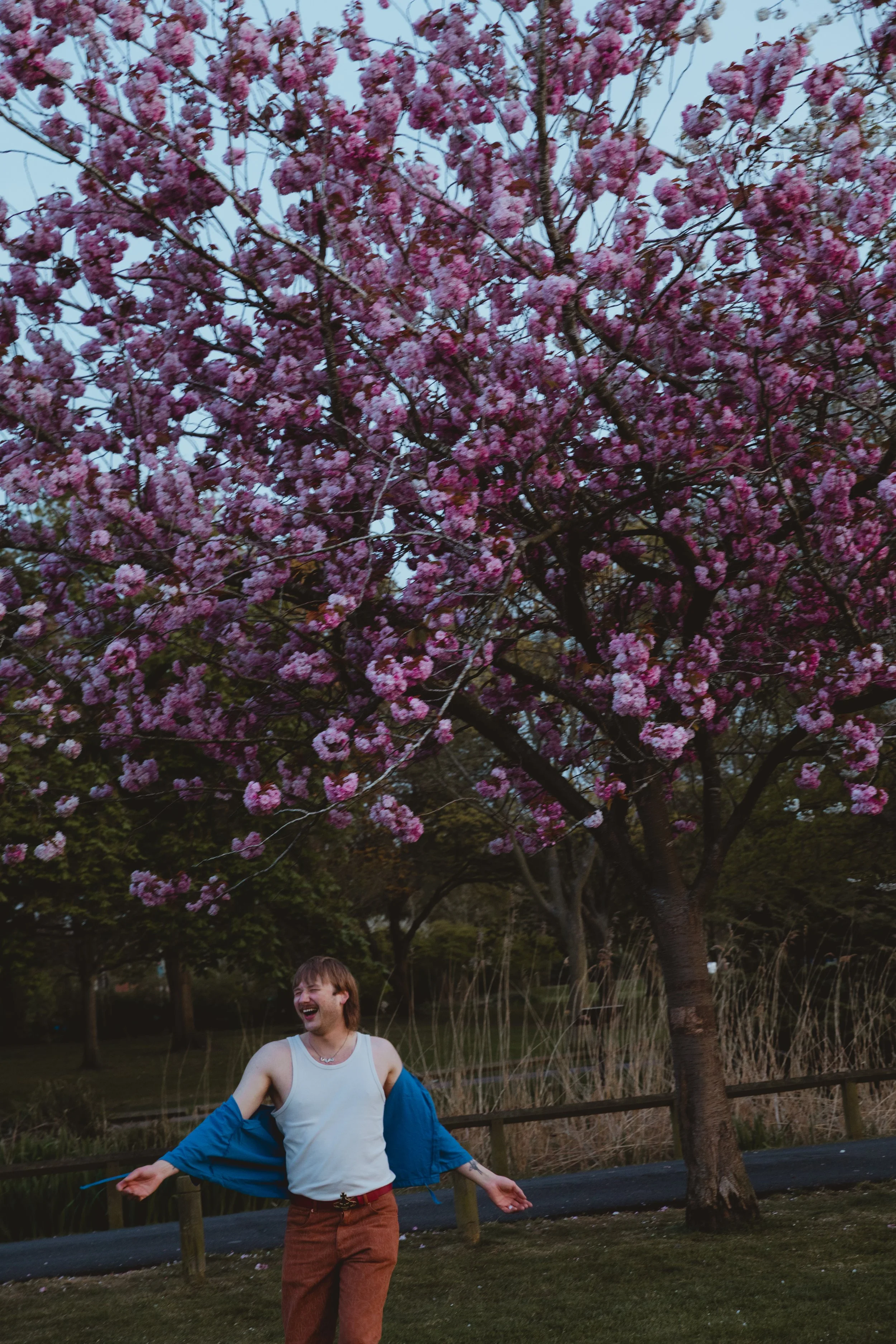 Portrait photograph of well dressed make yoga teacher with moustache in brown and blue outfit frolicking in a park in front of a pink blossom tree at Oulton Broad Suffolk 
