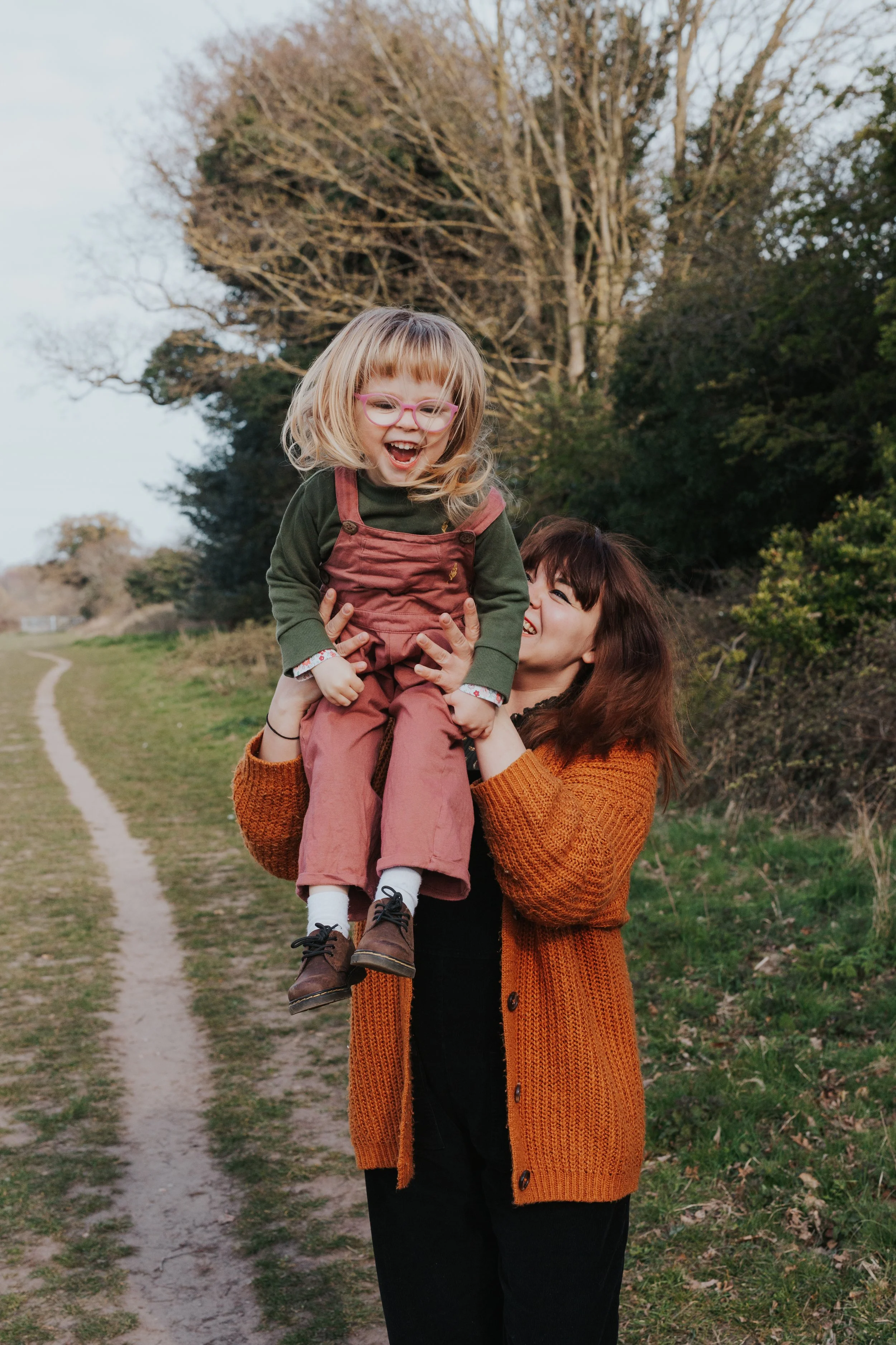 Mother in orange cardigan with daughter with blonde hair in green and pink outfit lifting the toddler into the air against a green backdrop taken at Lound Lakes Suffolk 
