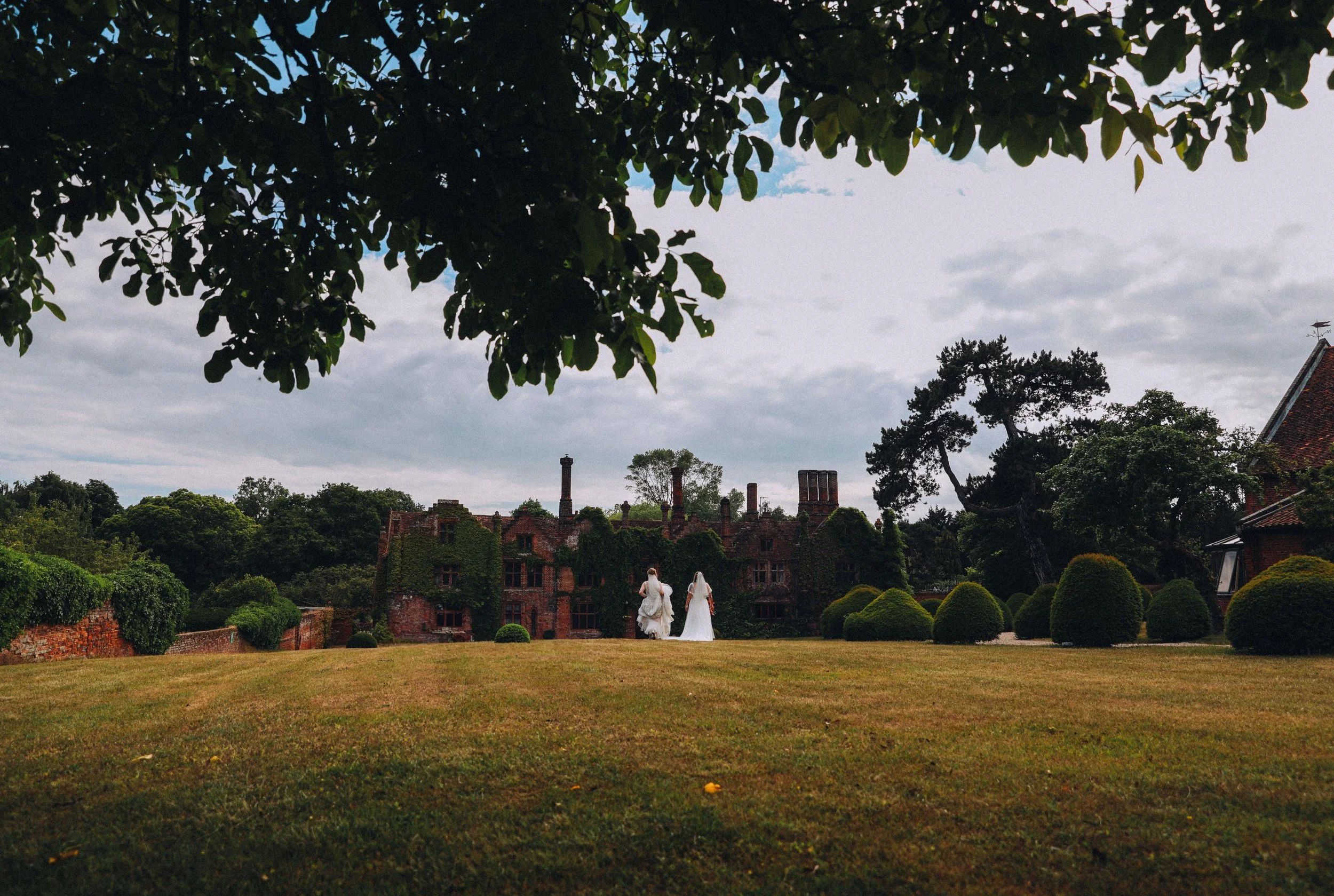 same sex couple bride and bride walking off in the distance across a manicured lawn against the backdrop of a luxury tudor mansion Ipswich Suffolk Seckford Hall Hotel and Spa 