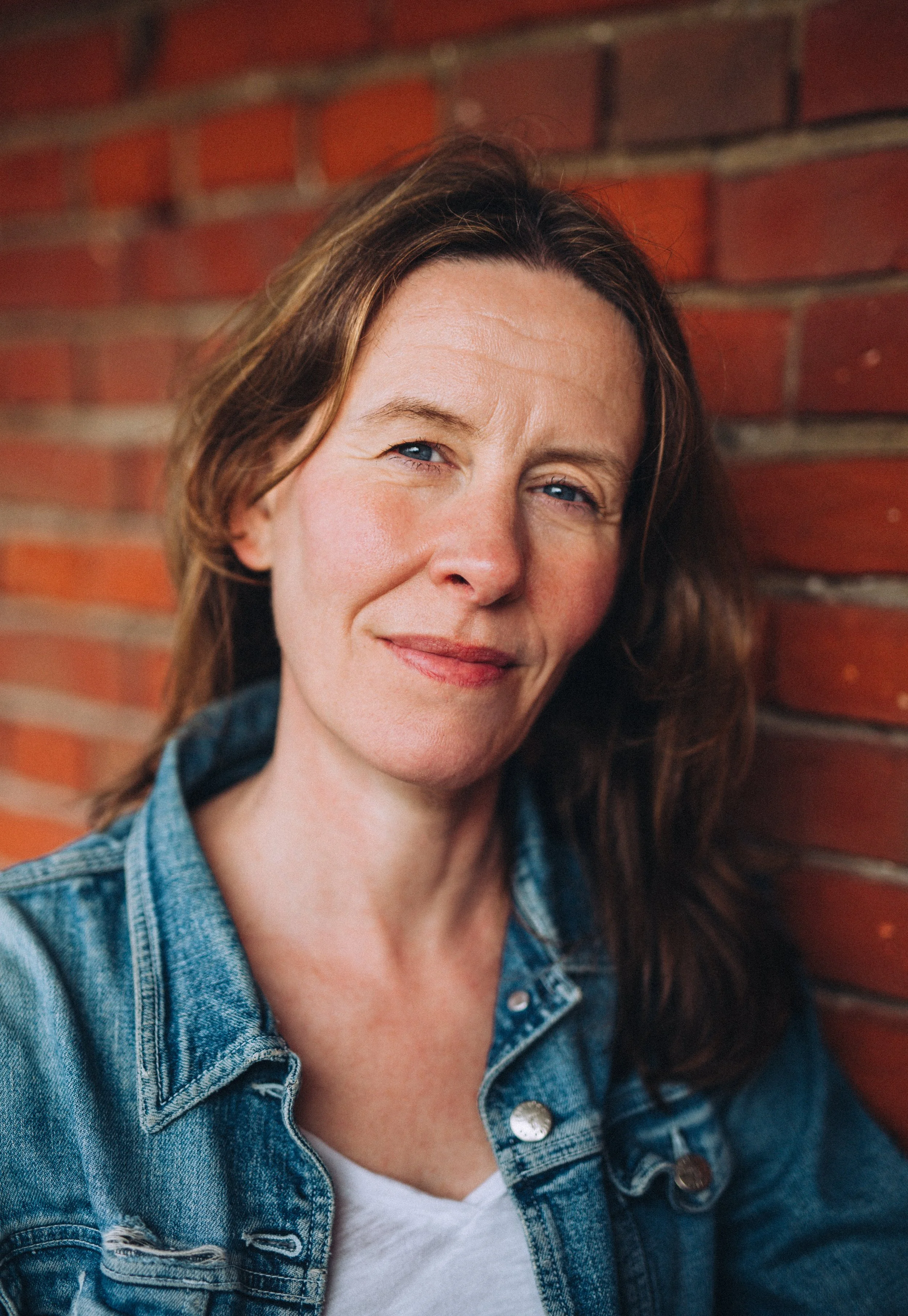 Softly lit portrait of an actor woman resting against a brick backdrop looking directly at the camera with a soft smile and denim jacket in Cromer Norfolk 