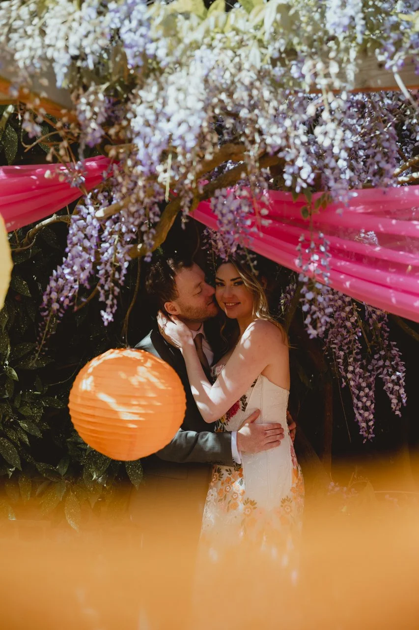 Vibrant editorial image of young bride in White Reflections boho dress and groom under wisteria with pink draped and orange lanterns bohemian garden party gardens at Chaucer Barn Holt Gresham Norfolk 
