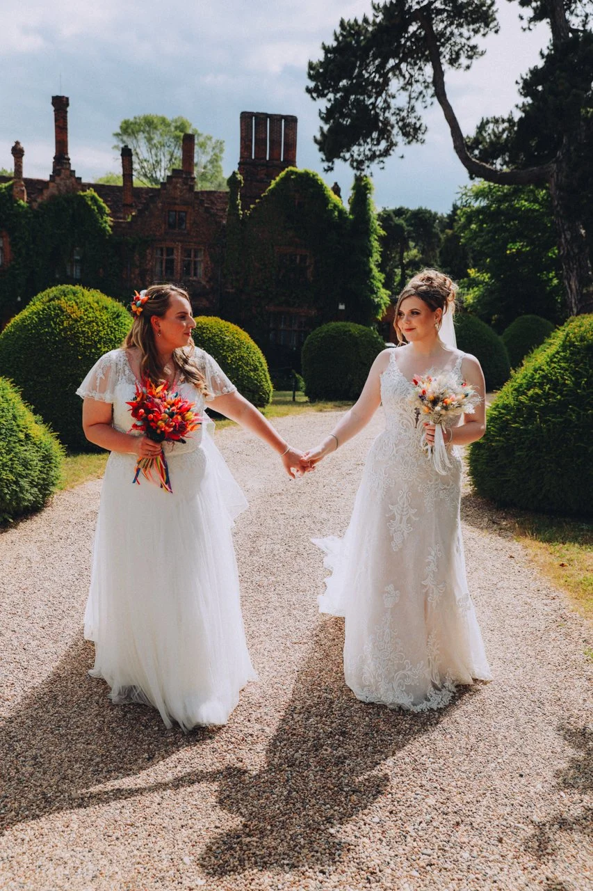 Same sex bride couple looking at each other in brides dresses holding hands with colourful dried flowers in a luxury topiary garden against the tudor mansion Seckford Hall Ipswich in the background 