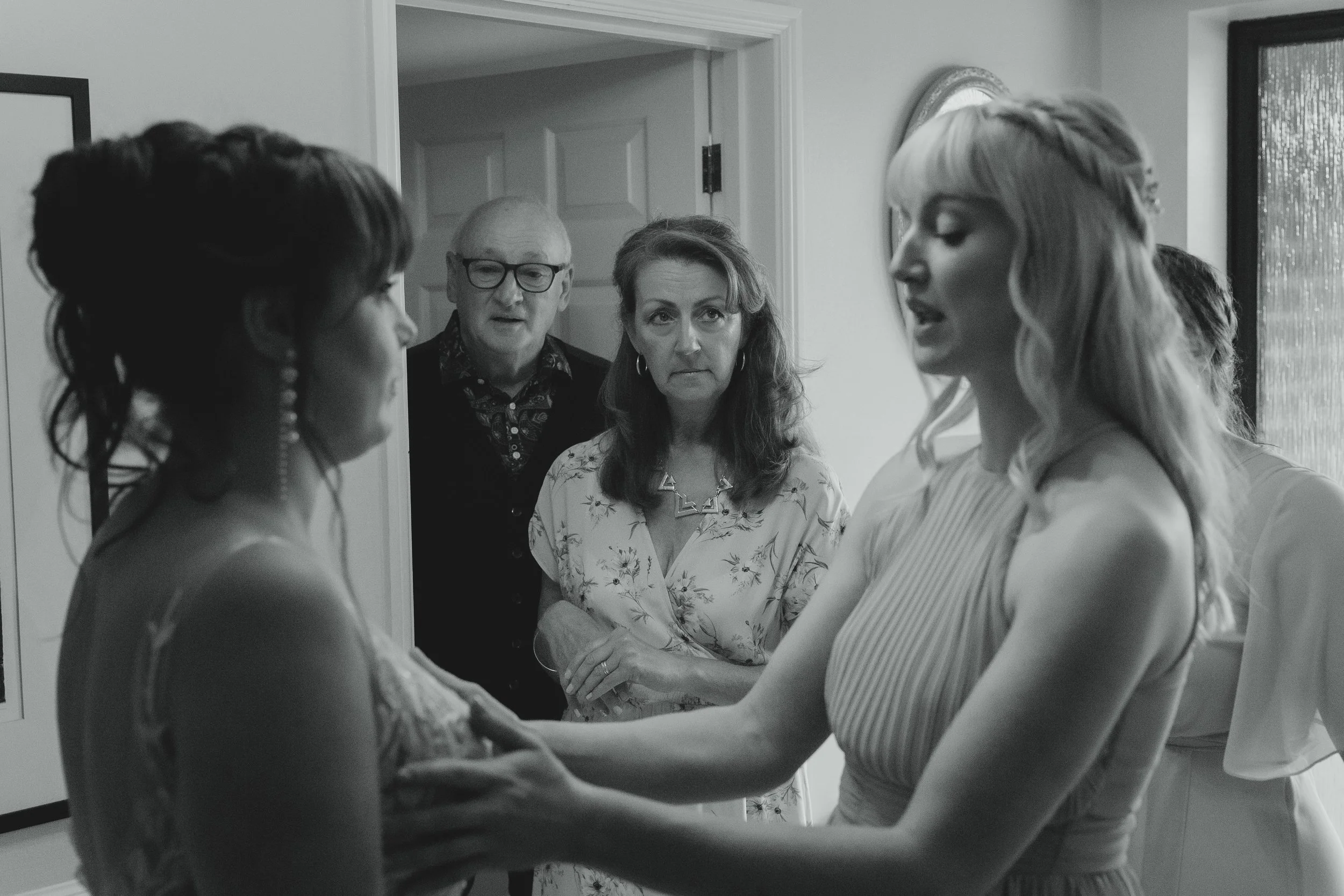 Black and white documentary image of bridesmaid adjusting brides dress whilst mother and father of the bride look on indoors bridal prep Norfolk 