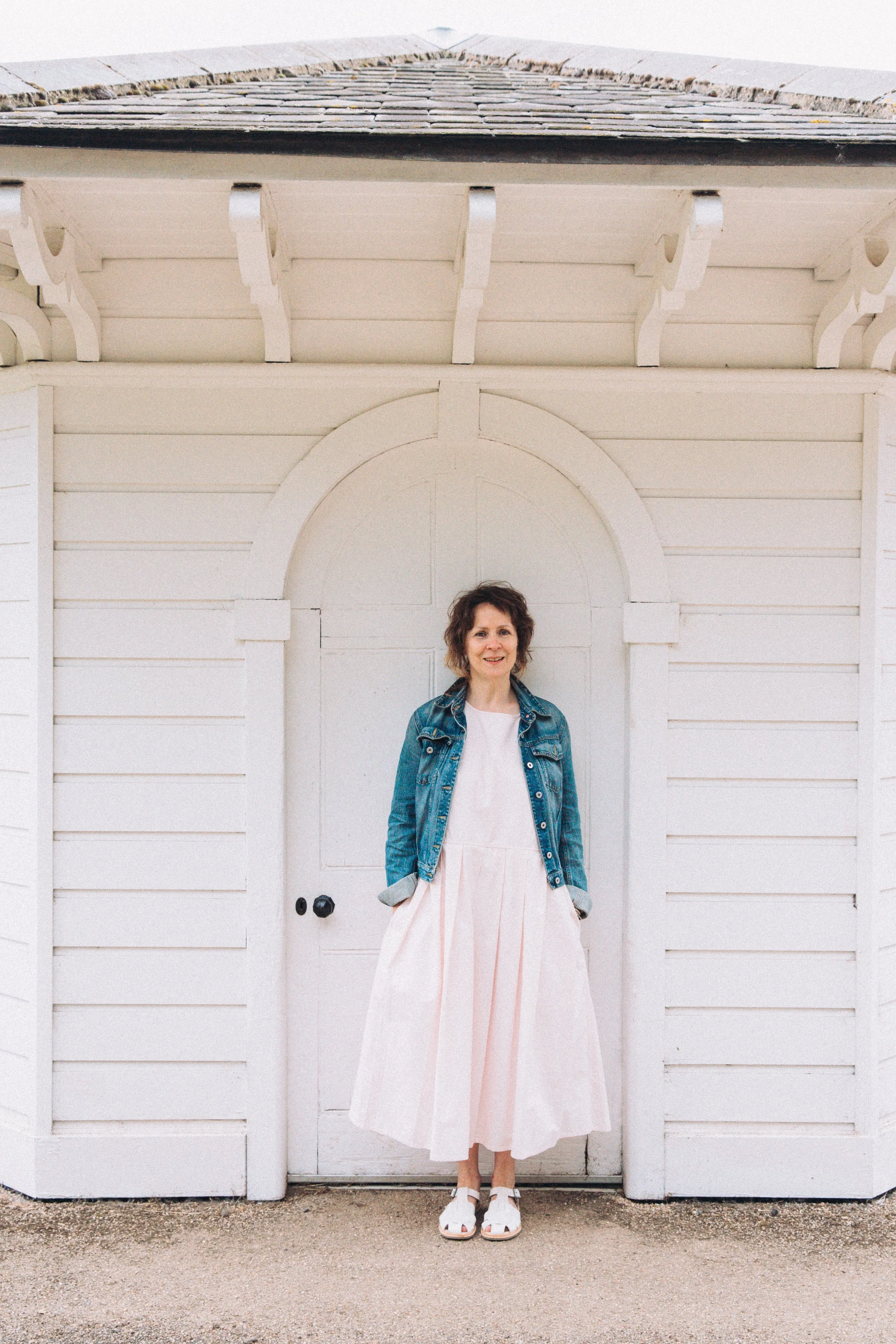 Portrait of a woman with dark hair and a white dress and denim jacket stood against a white clapboard building at Stowmarket Food Museum Suffolk 