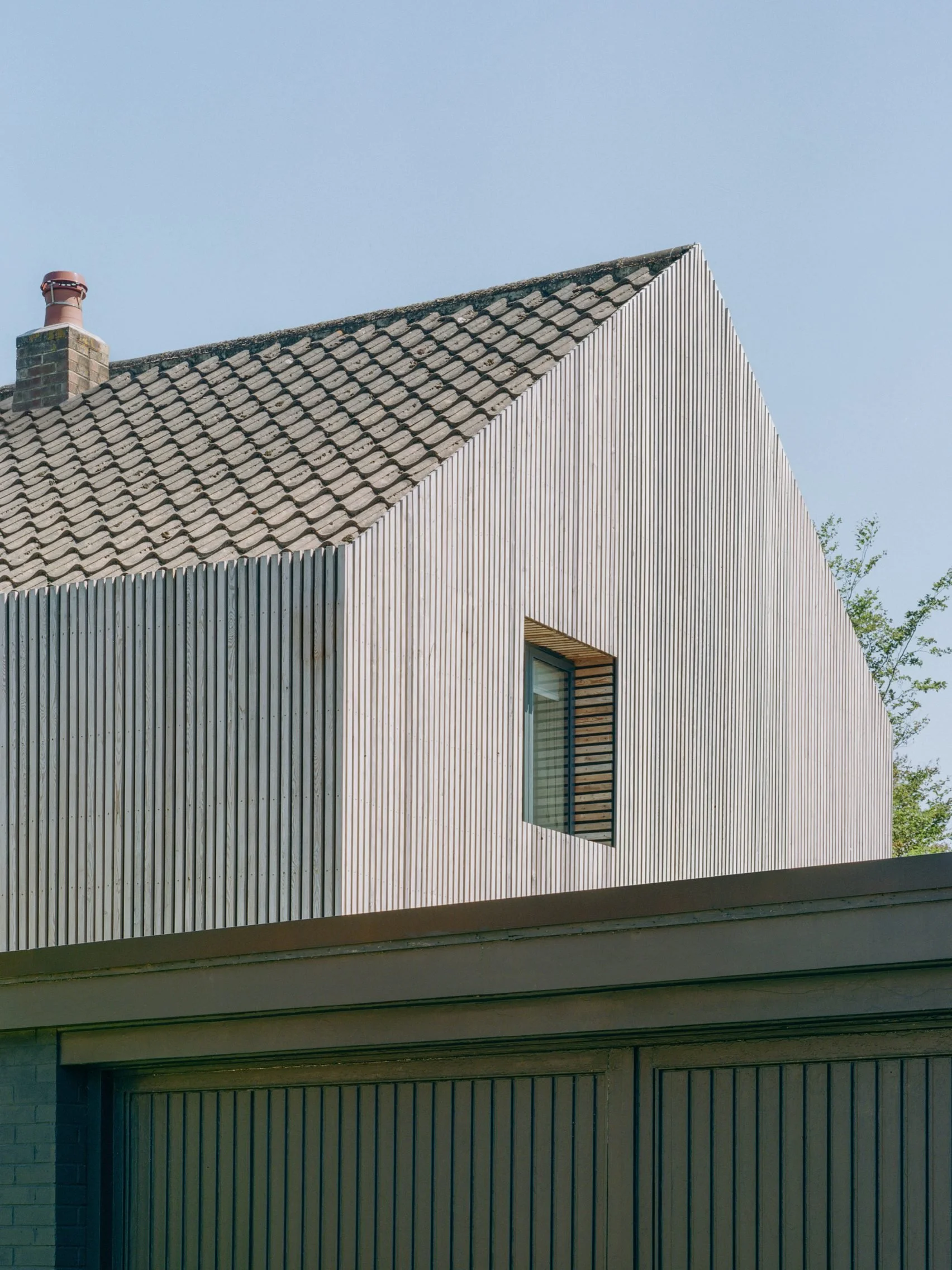 Modern house's upper floor with vertical wooden siding and a sloped roof with traditional shingles, a window with blinds, and a cloudy sky in the background.