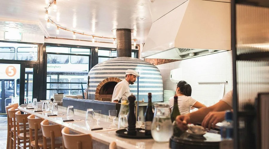 A restaurant interior with a long bar counter set with drinking glasses, bottles, and tableware. Two staff members are working behind the counter, with a chef in front of a wood-fired pizza oven. Large windows let in natural light.