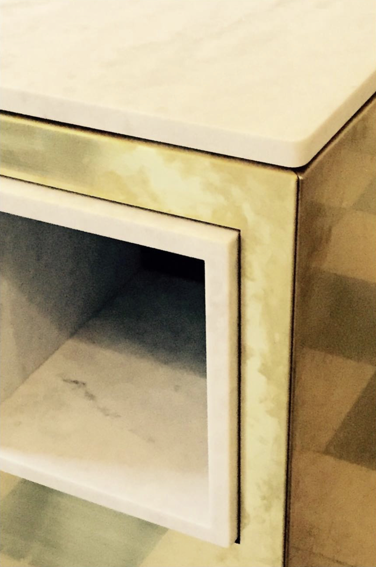 Close-up of a corner of a vintage nightstand or small cabinet, featuring gold and white painted finishes with some wear and patina, showing an open drawer with an interior that appears to be marble or marble-like material.