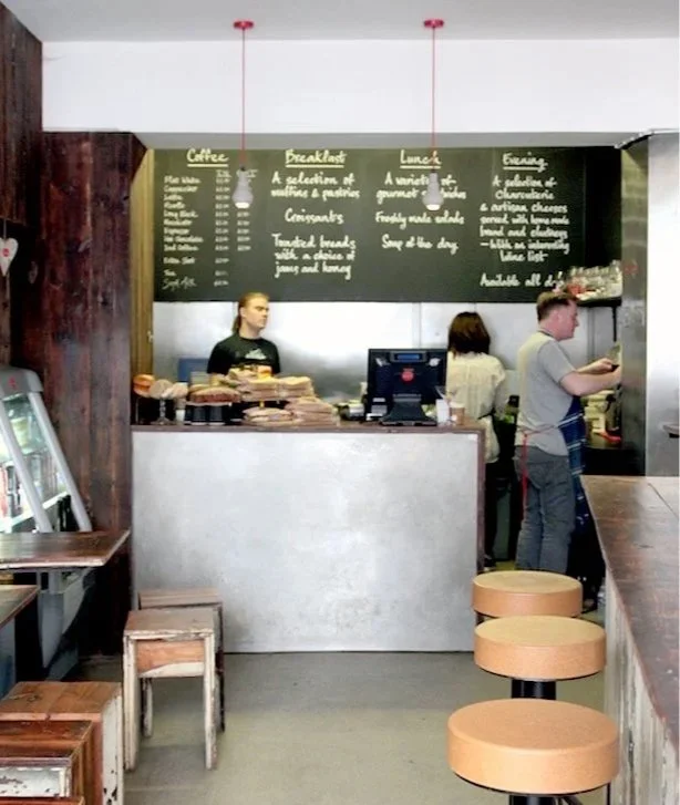 Interior of a cafe with a chalkboard menu, a counter with a cash register, and two customers ordering. There are stools on the right and small tables on the left.
