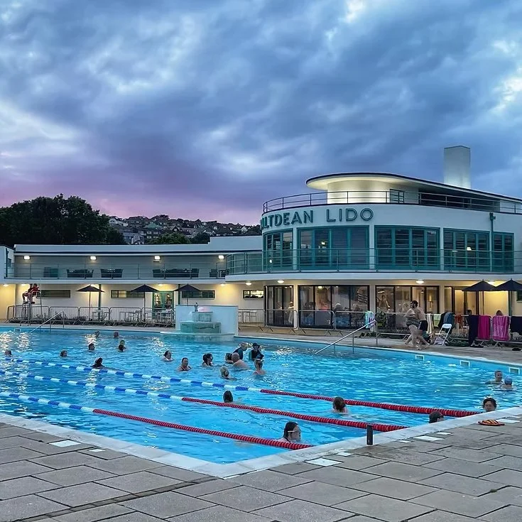 Outdoor swimming pool at sunset with people swimming and relaxing, surrounded by lounge chairs and umbrellas, with a modern building labeled 'OLDEAN LIDO' in the background.
