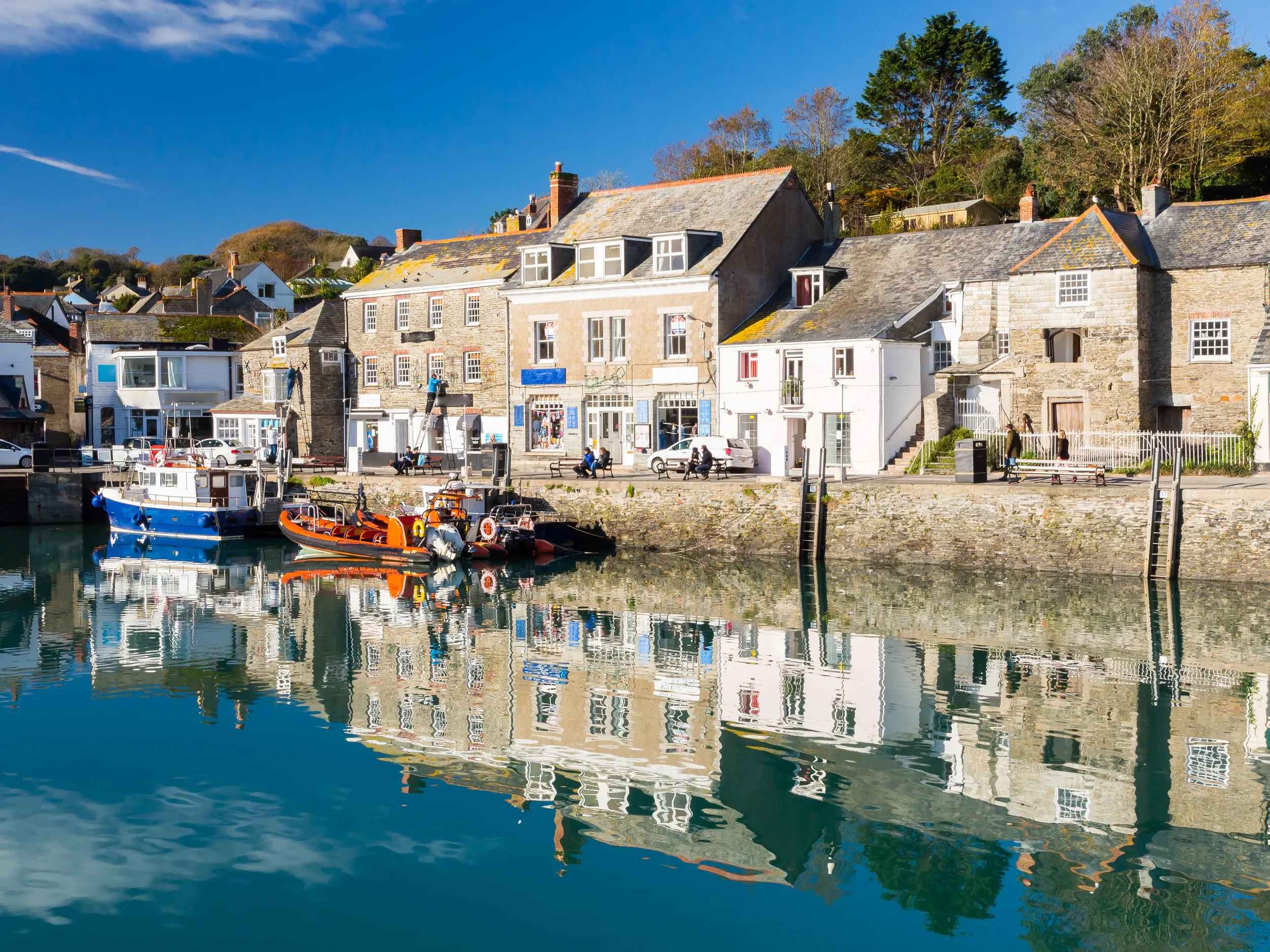A harbor scene with boats docked at the pier and colorful buildings along the waterfront, with trees and houses on a hill in the background under a blue sky.