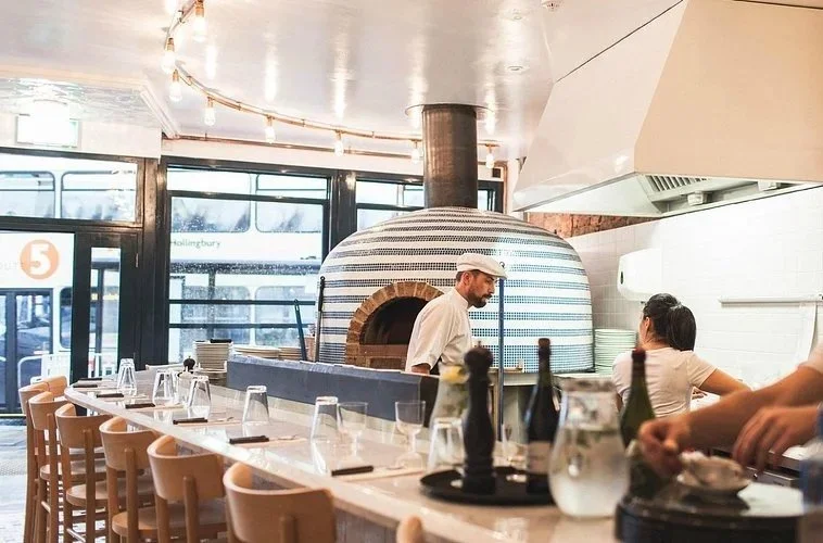 A chef baking in a pizza oven inside a restaurant with a bar seating area in the foreground, where glasses and bottles are arranged on the counter.