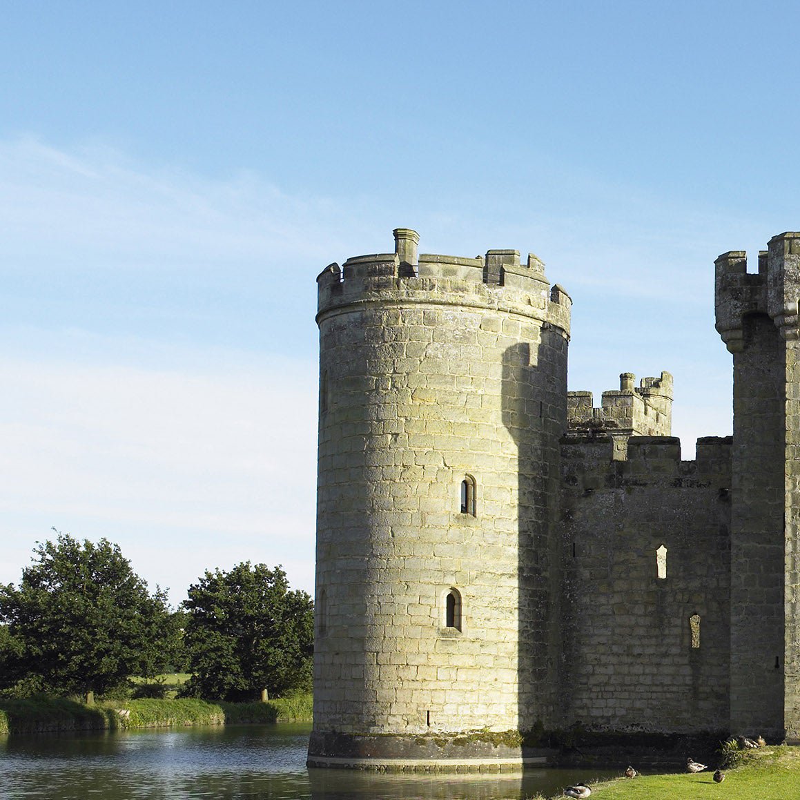 A stone castle tower with battlements, surrounded by water and trees under a blue sky.