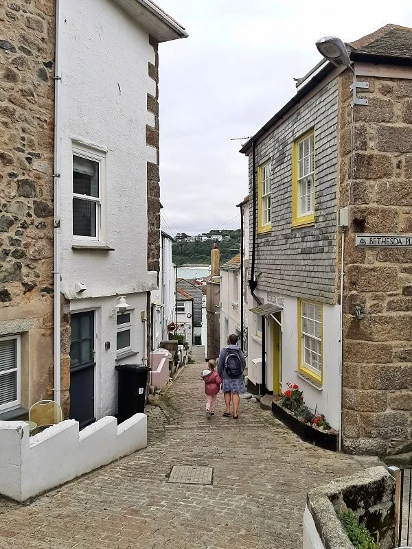 Two people, an adult and a child, walking down a narrow cobblestone street with houses on either side, overlooking a body of water in the distance.