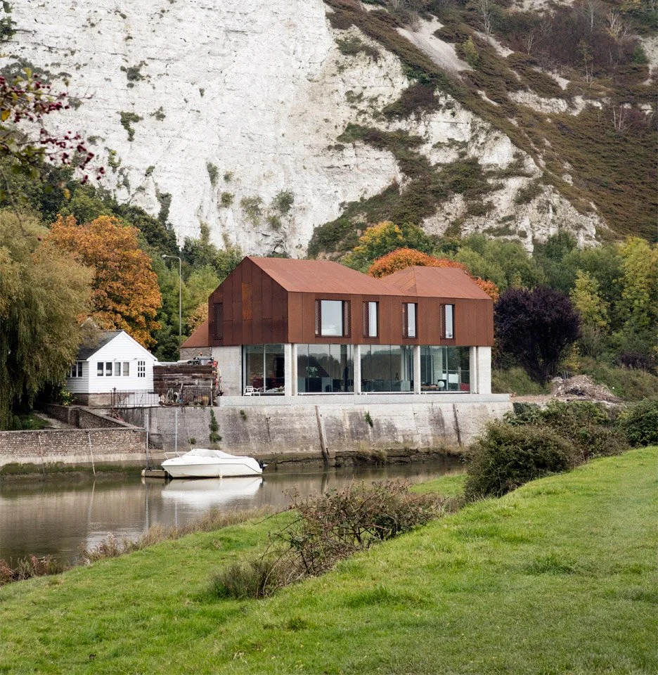 Modern house with large glass windows and rust-colored metal exterior, situated by a river with a small boat, surrounded by trees and hills.