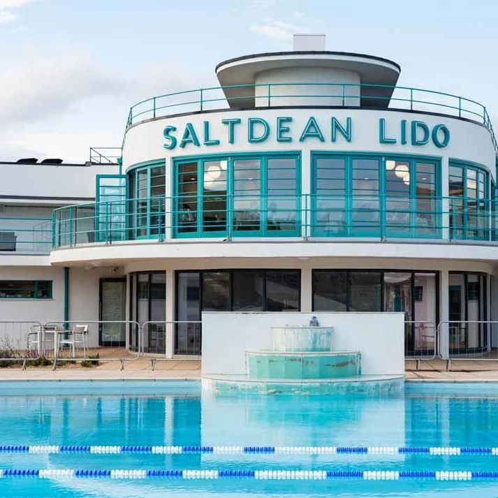 Modern swimming pool facility named Saltdean Lido with a circular upper deck and large windows, outdoor pool in foreground, and cloudy sky.