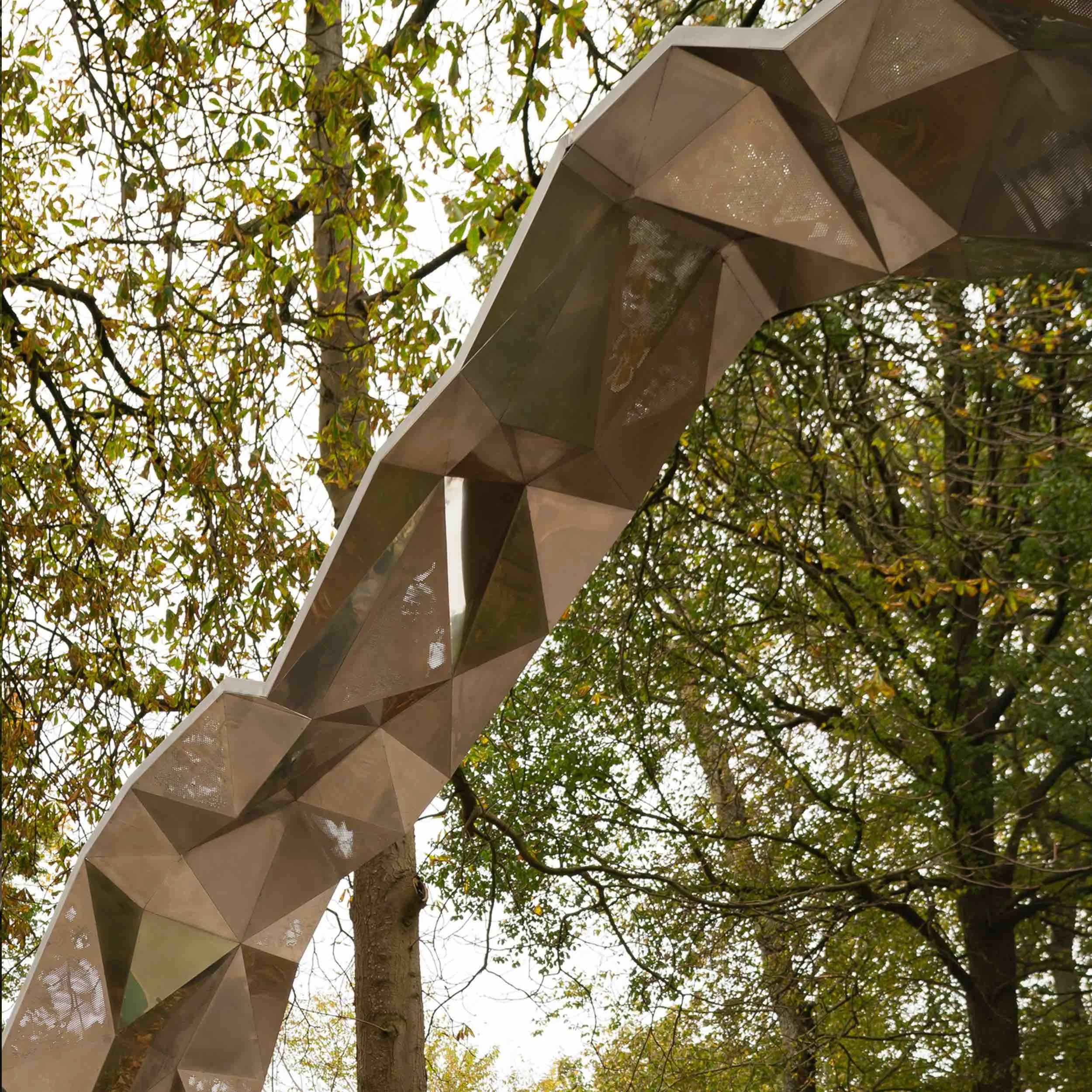 A geometric metal sculpture running through a tree canopy with green leaves, viewed from below.
