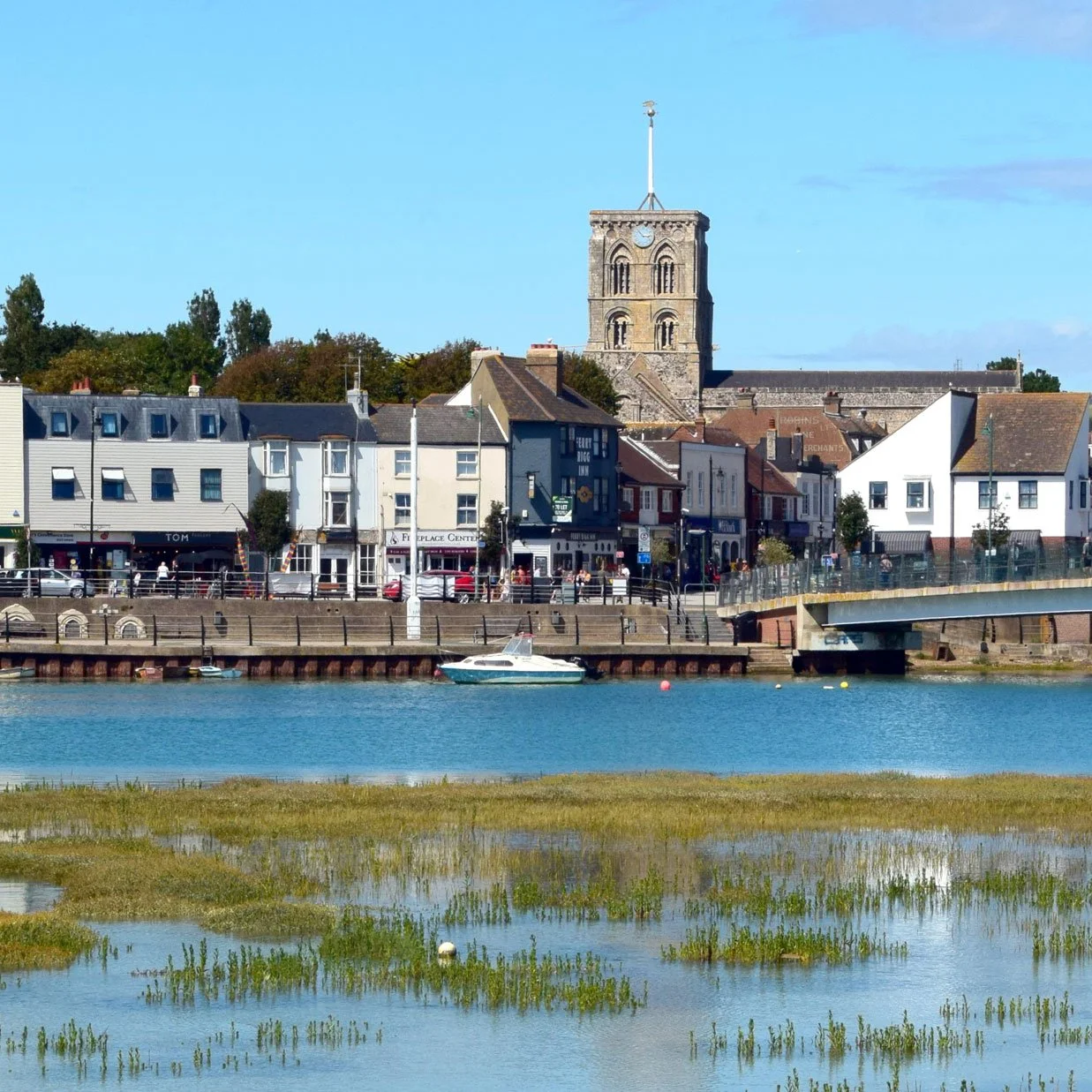 A scenic waterfront view of a small town with colorful buildings, a church with a tall clock tower, a boat in the water, and a grassy marsh in the foreground.