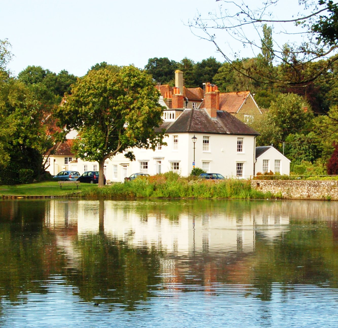A white house with multiple chimneys and a slate roof sits beside a body of water, surrounded by green trees and foliage, with a grassy lawn and a stone wall in front.