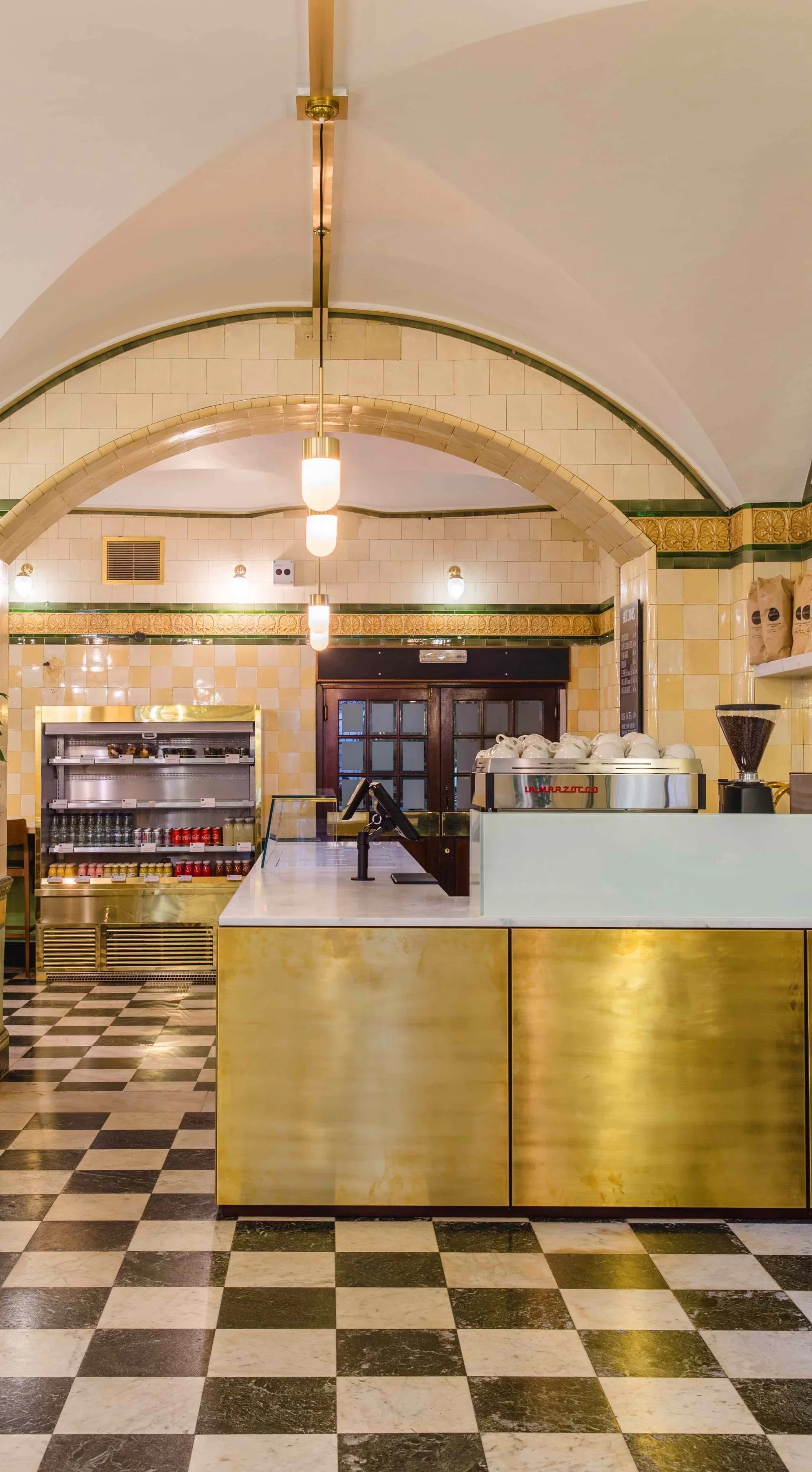 Interior of a vintage cafe or bakery with checkered black and white floor tiles, a golden counter, and a tiled archway ceiling with pendant and wall lights, a coffee machine, and shelves with jars.