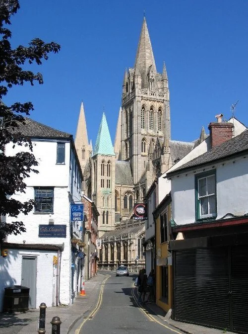 View of a tall Gothic cathedral with pointed spires, seen from a narrow street lined with small shops and buildings with colorful facades.