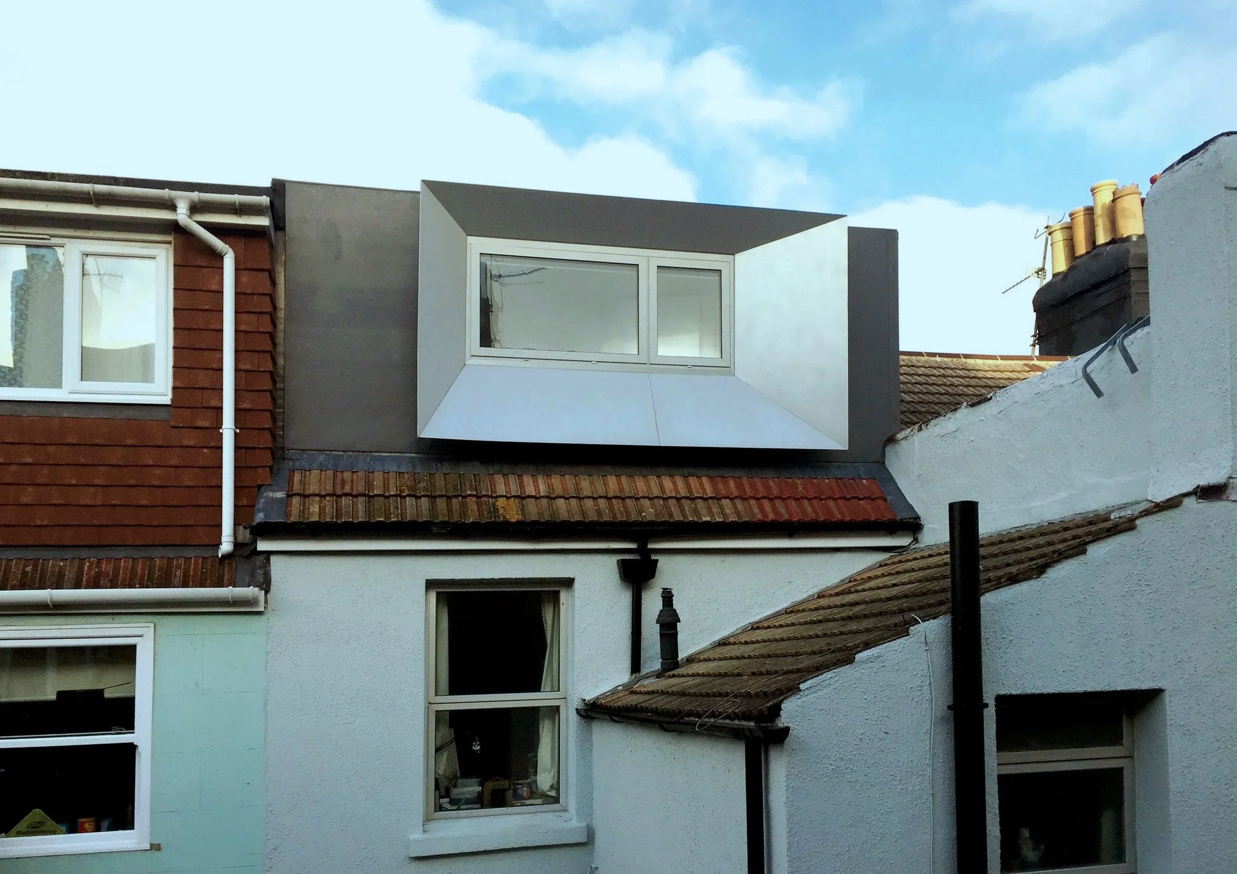 A residential building exterior showing a white painted house with multiple windows, a red-tiled roof, and an added modern dormer window with a metallic surround.