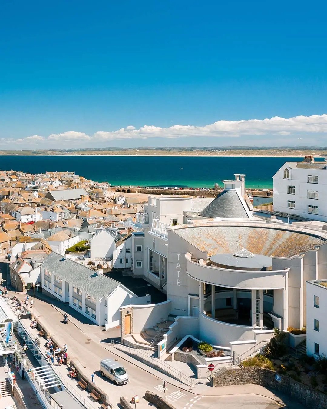 A coastal town with white buildings and a large body of water under a bright blue sky with some clouds.