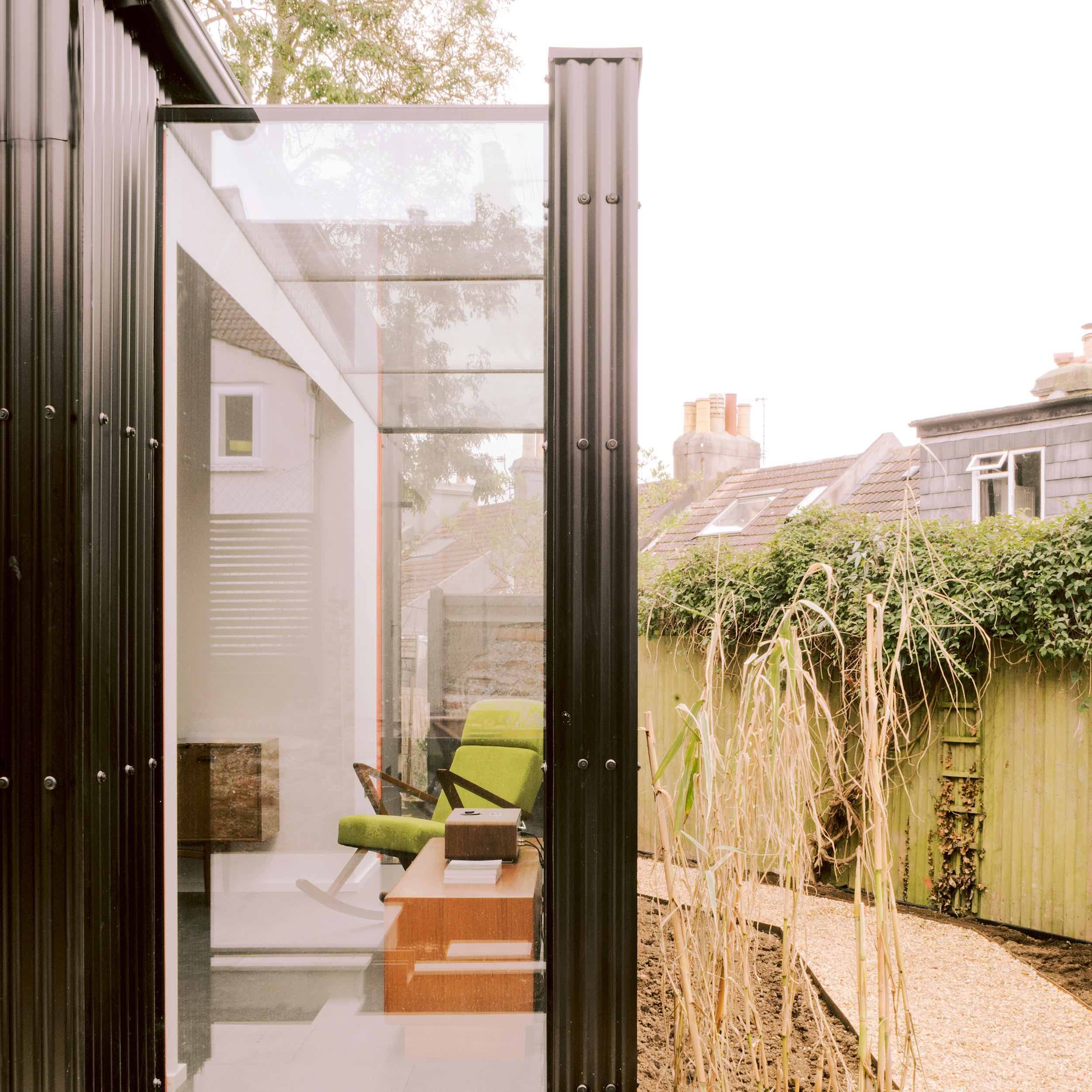 View through a glass door into a home office with a green chair, bookshelf, and window, overlooking a garden with dried plants and a wooden fence.