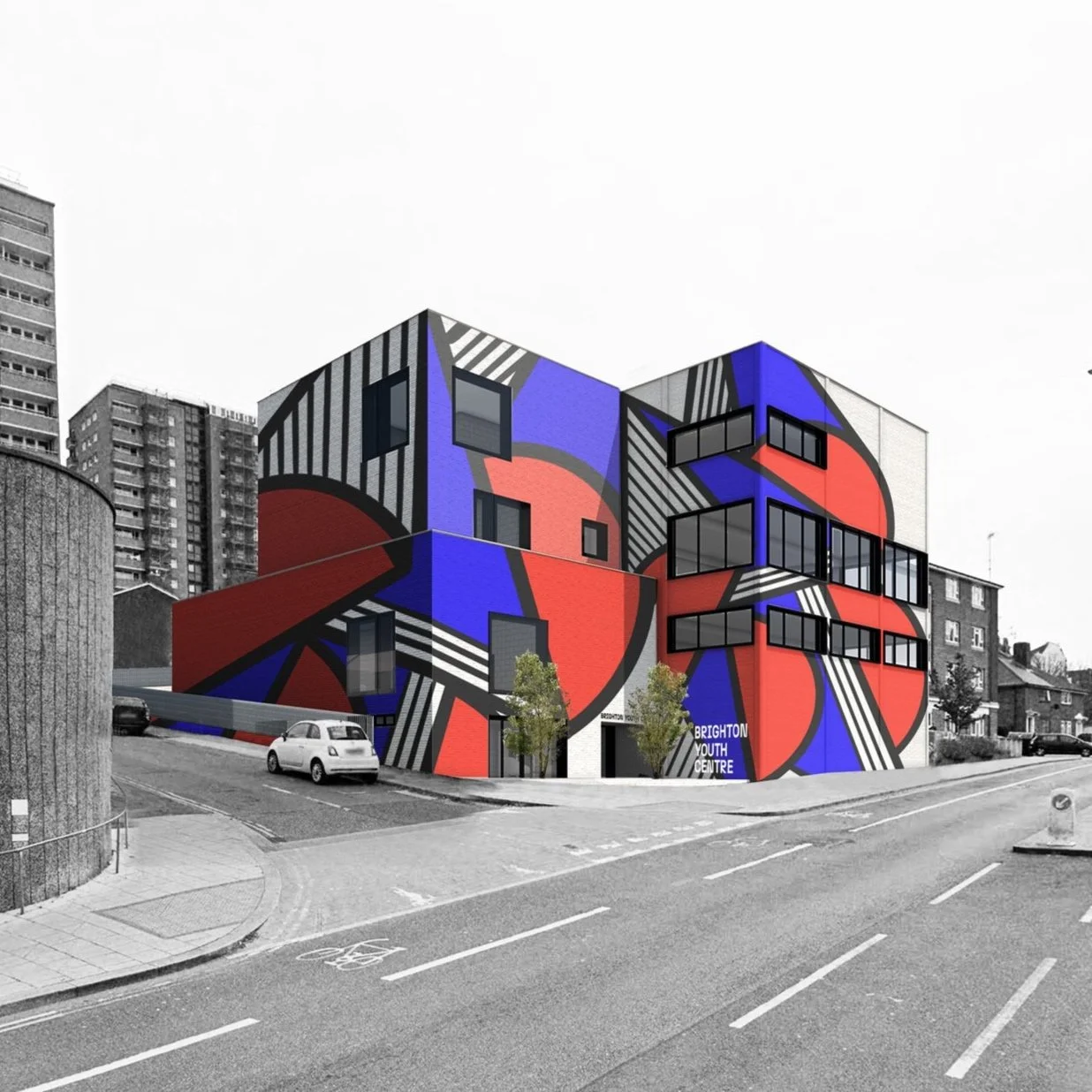 A modern building with a colorful mural featuring geometric shapes in red, blue, black, and white. The building has multiple floors with large windows, located on a street corner with parked cars and small trees in front. In the background, there are taller residential buildings.