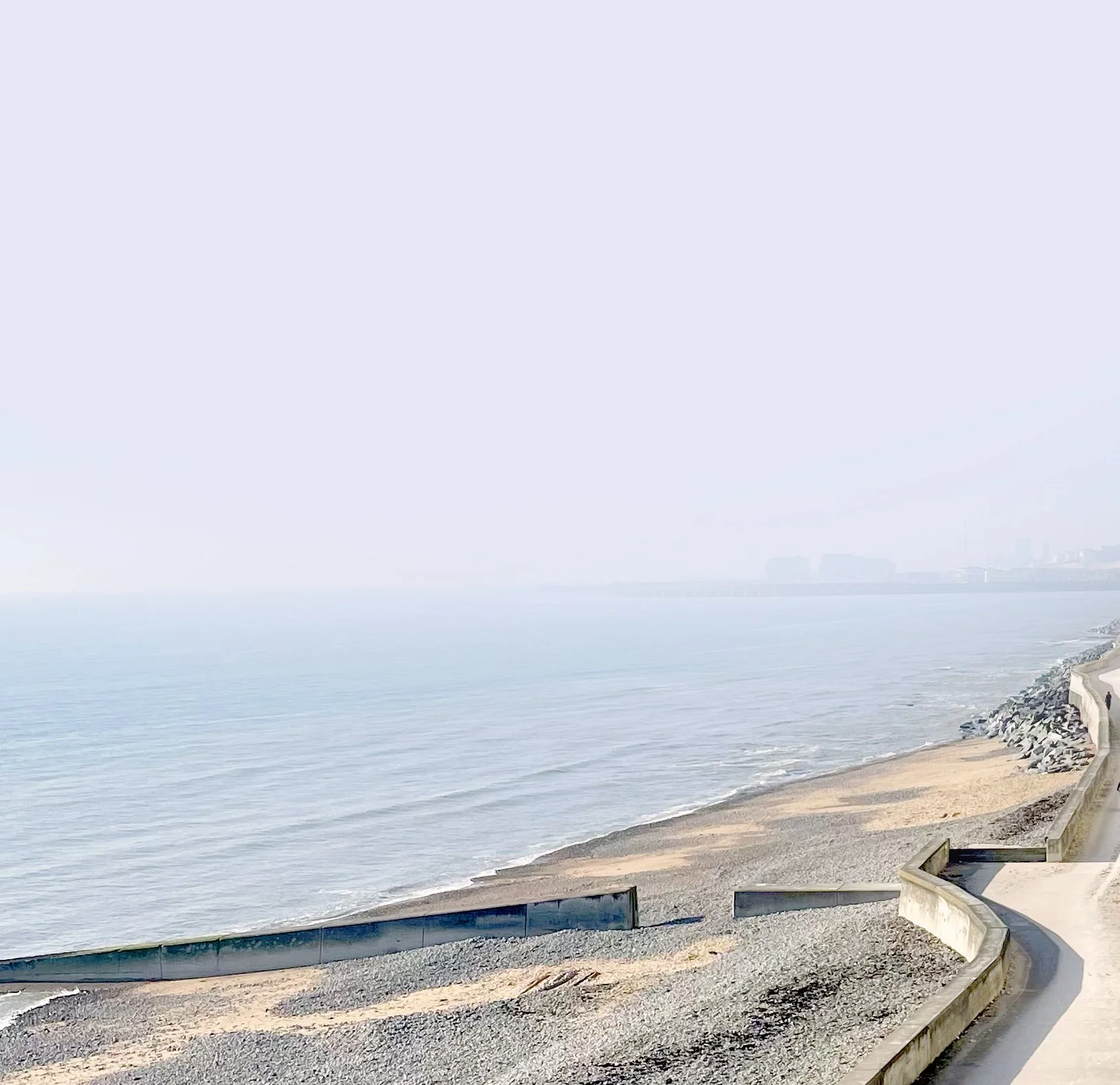 A shoreline with a walking path and a curved concrete barrier, with rocks along the beach and water extending into the horizon. There are distant buildings faintly visible through haze.