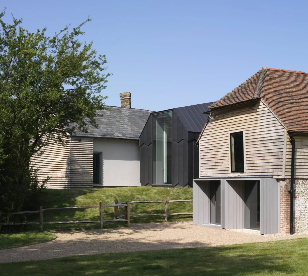 A modern house with a mix of traditional and contemporary architectural styles, featuring wooden, brick, and metal siding, a small grassy yard, and a blue sky.