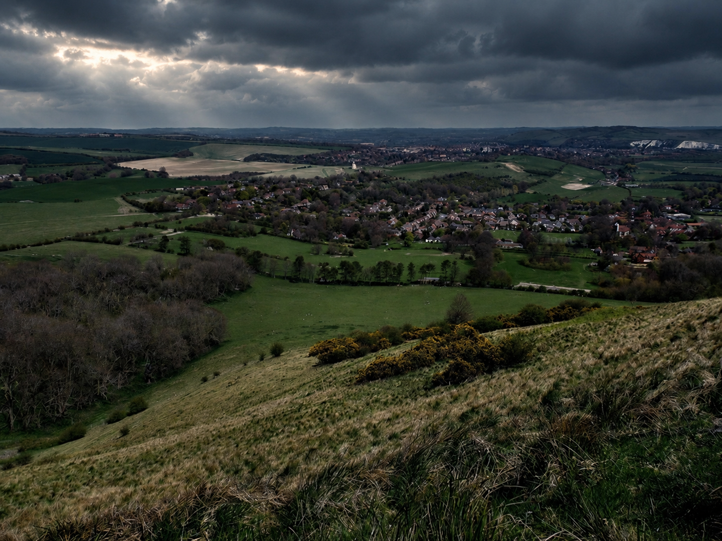 A landscape view of a rural area with houses, green fields, trees, and rolling hills under a cloudy sky.