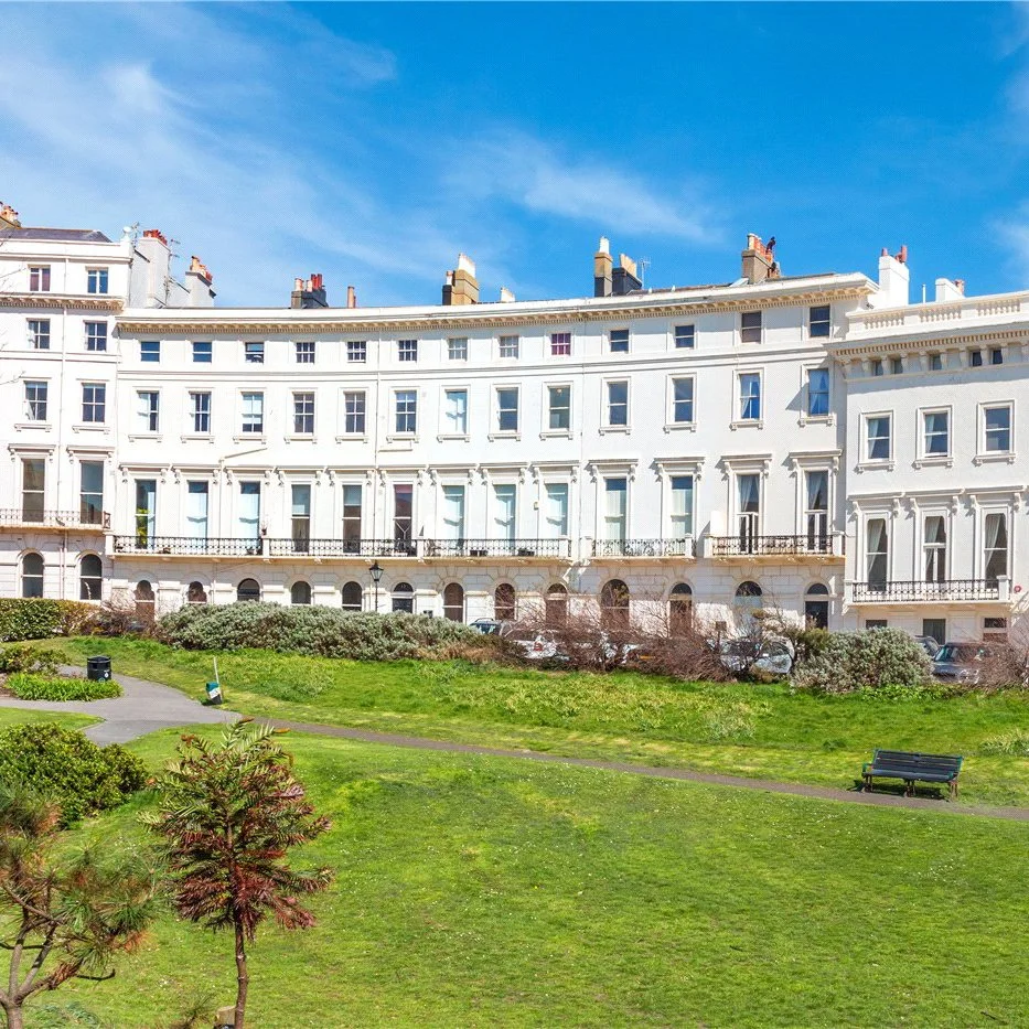 White Victorian-style apartment building with multiple windows, overlooking a grassy park with benches and walking paths under a blue sky.